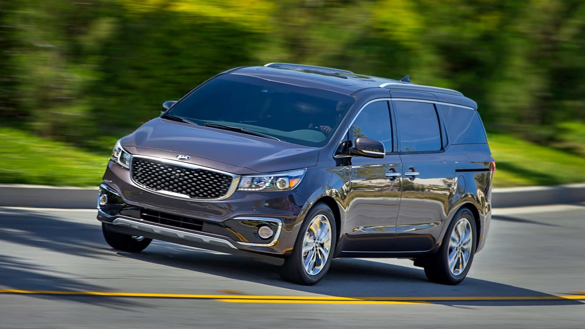 Kia Sedona 桌布 壁紙 — 一架時尚深灰色Kia小型客車在陽光明媚的道路上行駛，背景有模糊的綠色樹葉。
