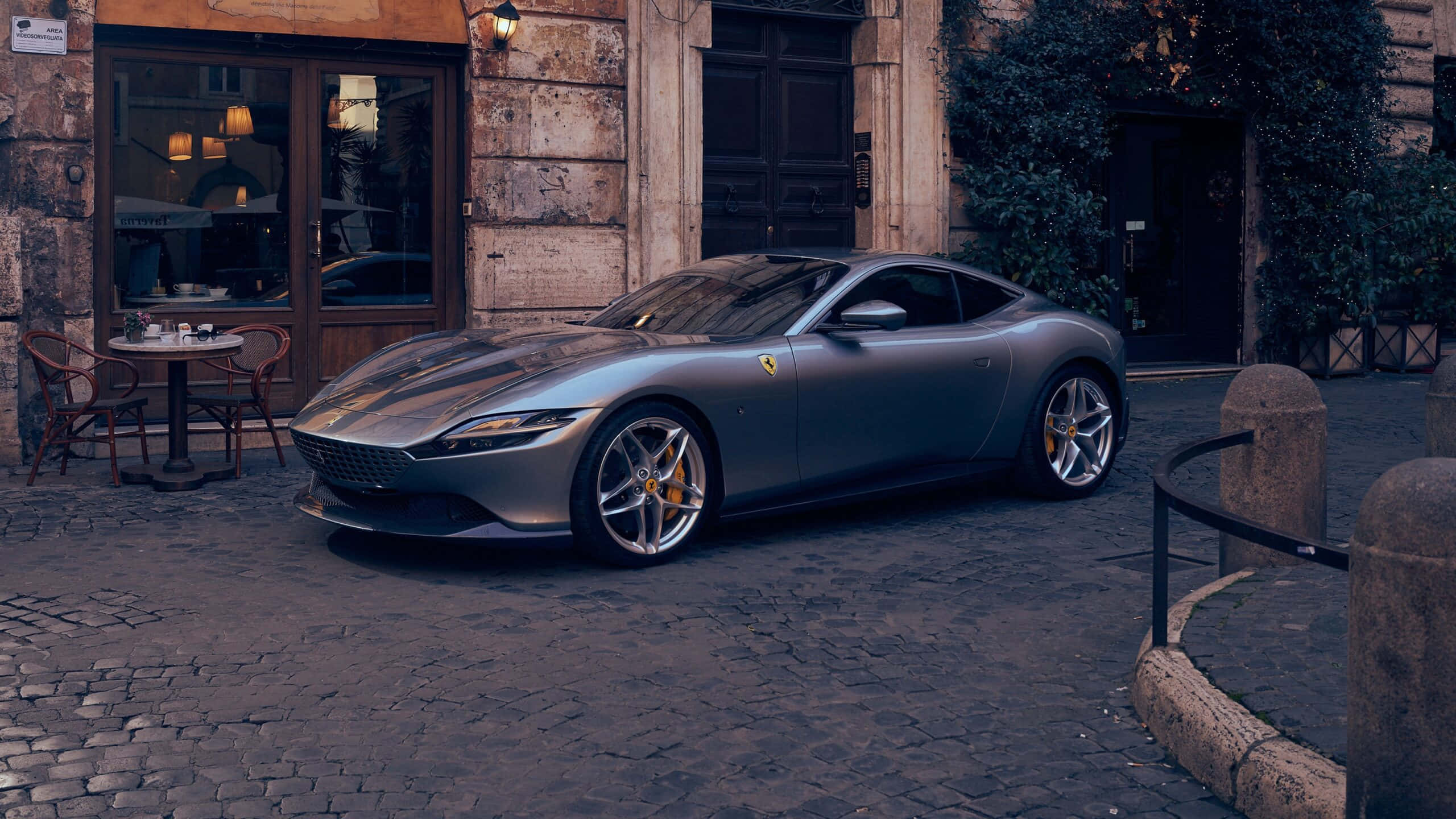 Ferrari Roma 桌布 壁紙 — 一架時尚的銀色法拉利跑車停泊在鵝卵石街道上，旁邊是一間帶有溫暖室內燈光的鄉村風格咖啡館。