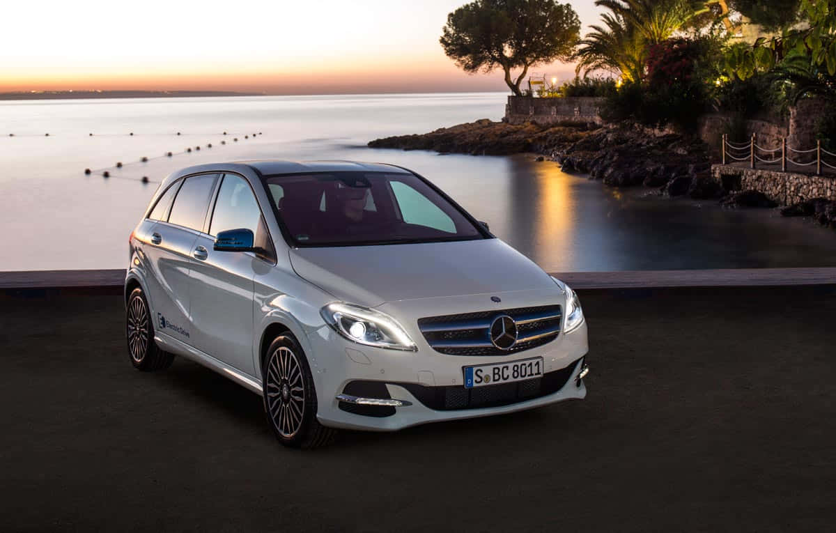 Mercedes Benz B-class 桌布 壁紙 — 一架時尚的白色 Mercedes-Benz B-Class 電動車停泊在日落時分的海濱長廊，背景有平靜的海水和棕櫚樹。
