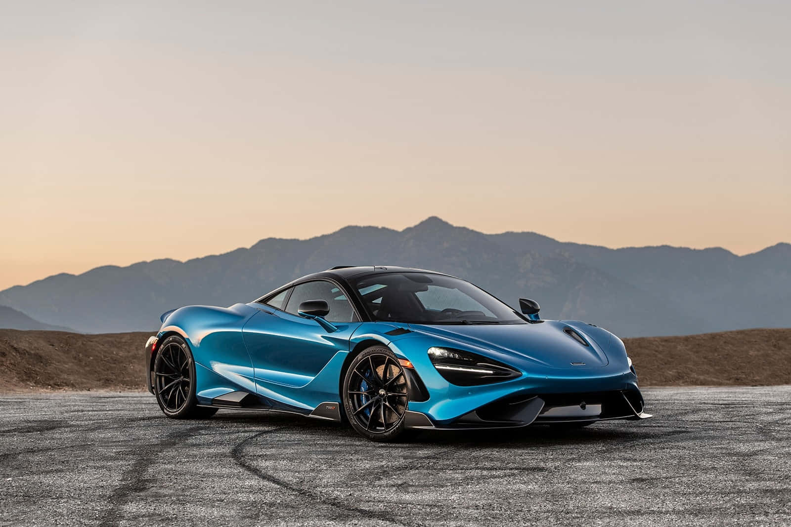 Mclaren 765lt 桌布 壁紙 — 一架時尚的藍色跑車帶有黑色點綴，停在鋪設的賽道上，背景是日落時分的山脈。
