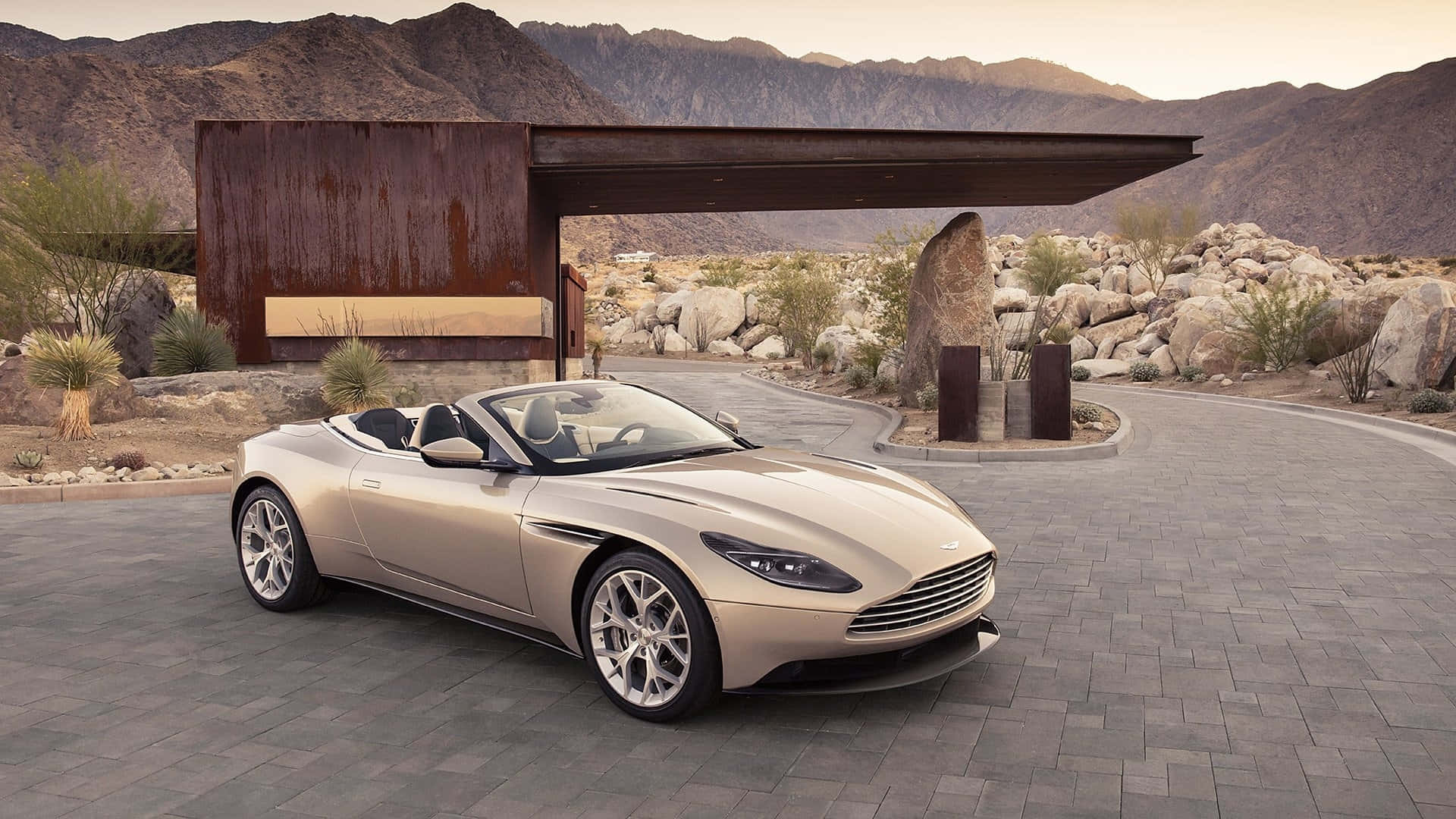Aston Martin Db11 桌布 壁紙 — 一架時尚的米色Aston Martin DB9開篷車停在石頭車道上，旁邊是現代生鏽的金屬結構，背景有山脈。