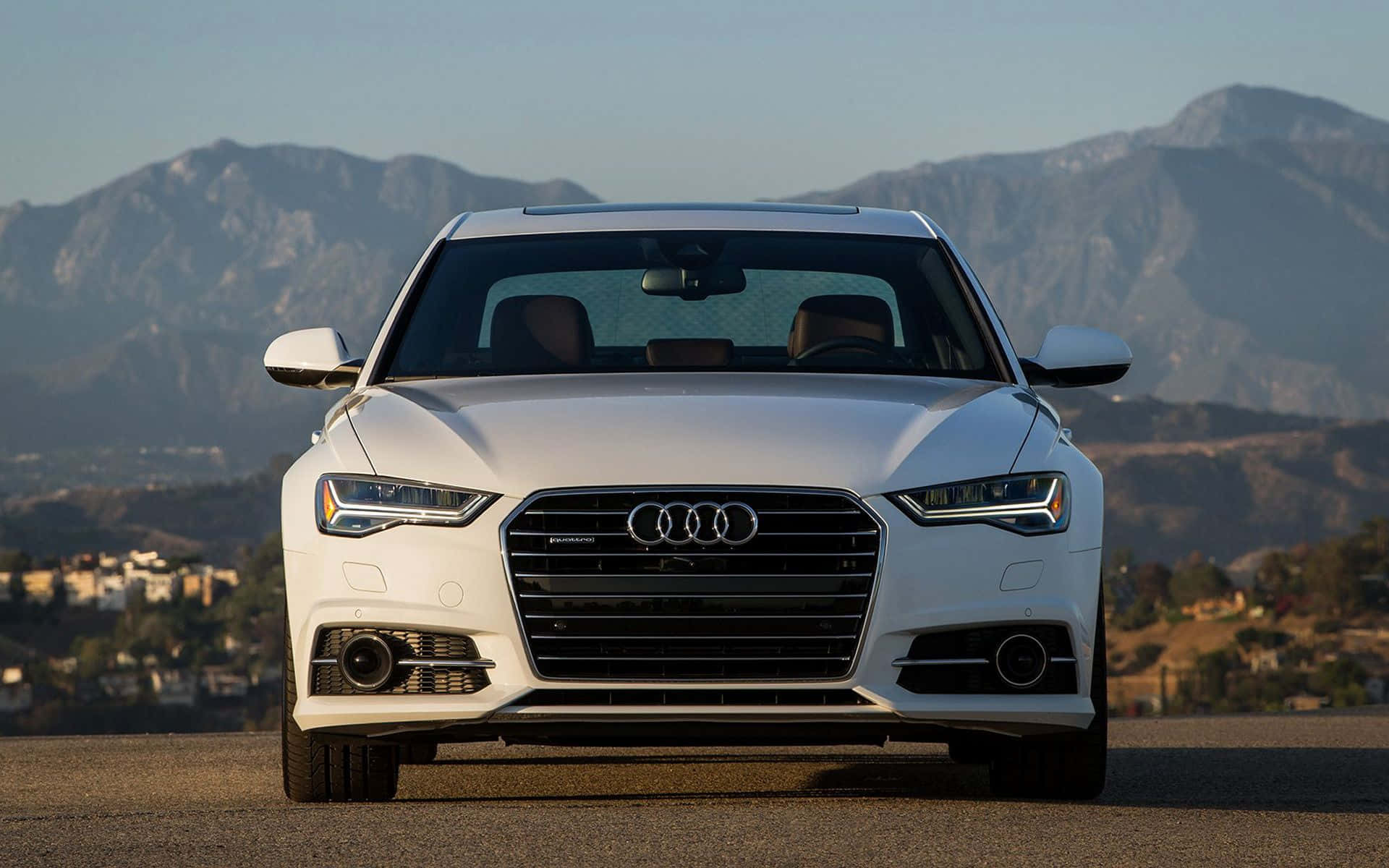 Audi A6 桌布 壁紙 — 一架時尚的白色奧迪轎車在鋪砌的道路上向前行駛，背景是崎嶇的山脈和晴朗的天空。