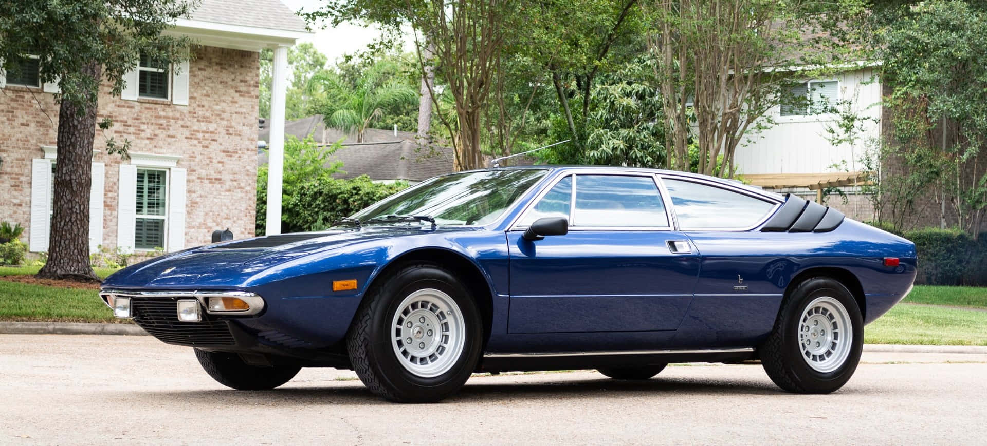 Lamborghini Urraco 桌布 壁紙 — 一架時尚的藍色經典跑車停泊在郊區街道上，背景有樹木和房屋。