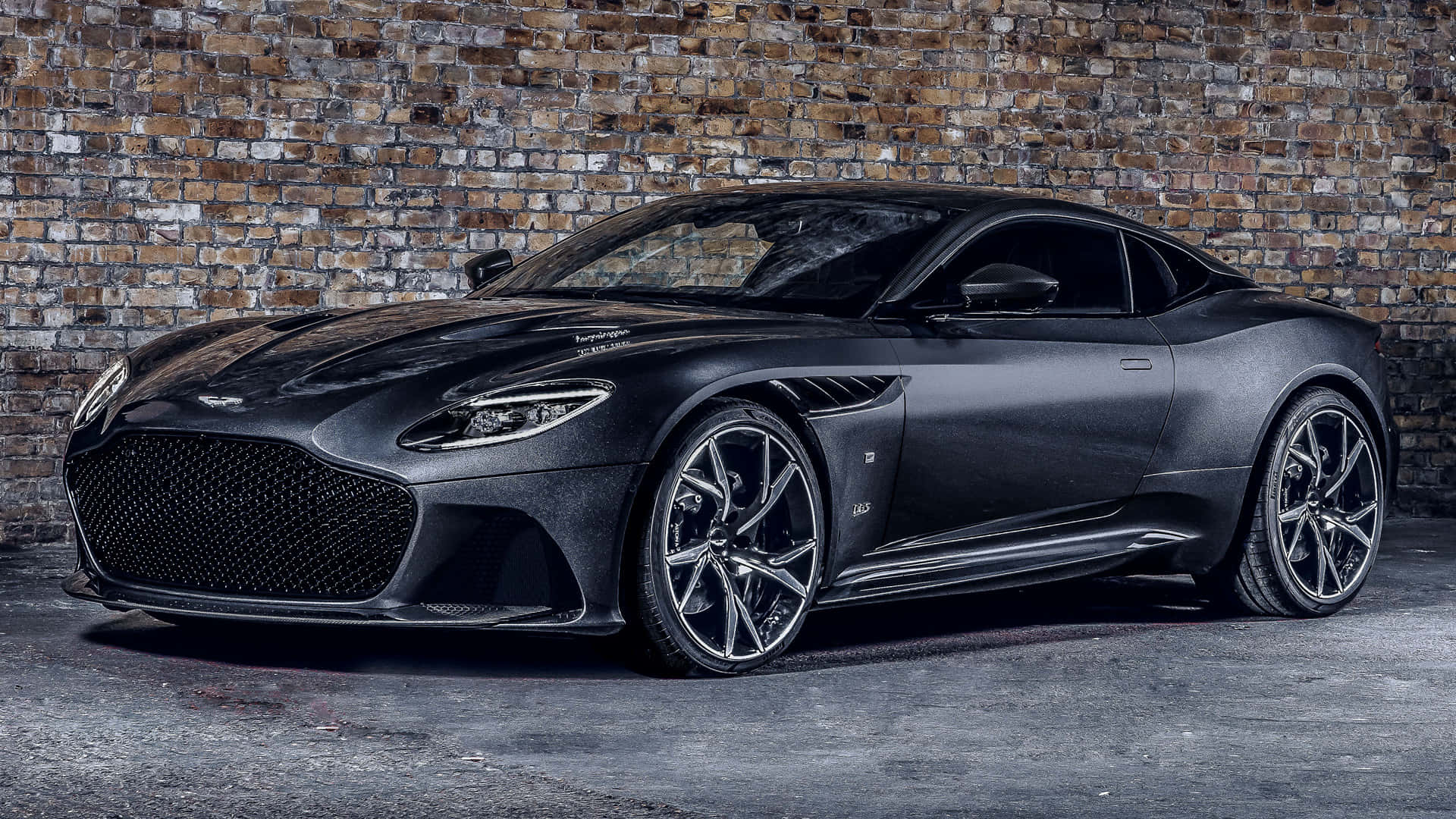 Aston Martin Dbs Superleggera 桌布 壁紙 — 一架時尚的啞光黑色 Aston Martin DBS Superleggera 停泊在昏暗車庫中風化的磚牆旁。