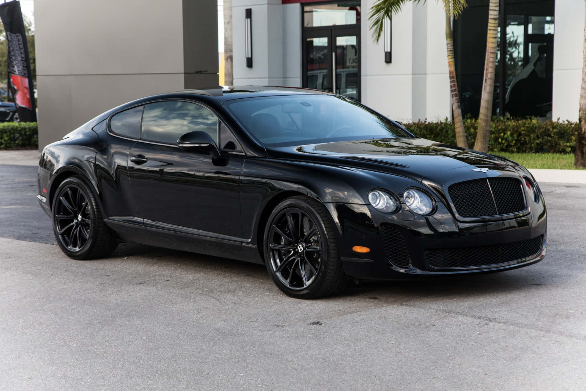 Bentley Continental Supersports 桌布 壁紙 — 一架時尚的黑色賓利 Continental GT 雙門轎跑車在日光下閃閃發光，停泊在鋪砌的停車場上，旁邊是現代建築和棕櫚樹。