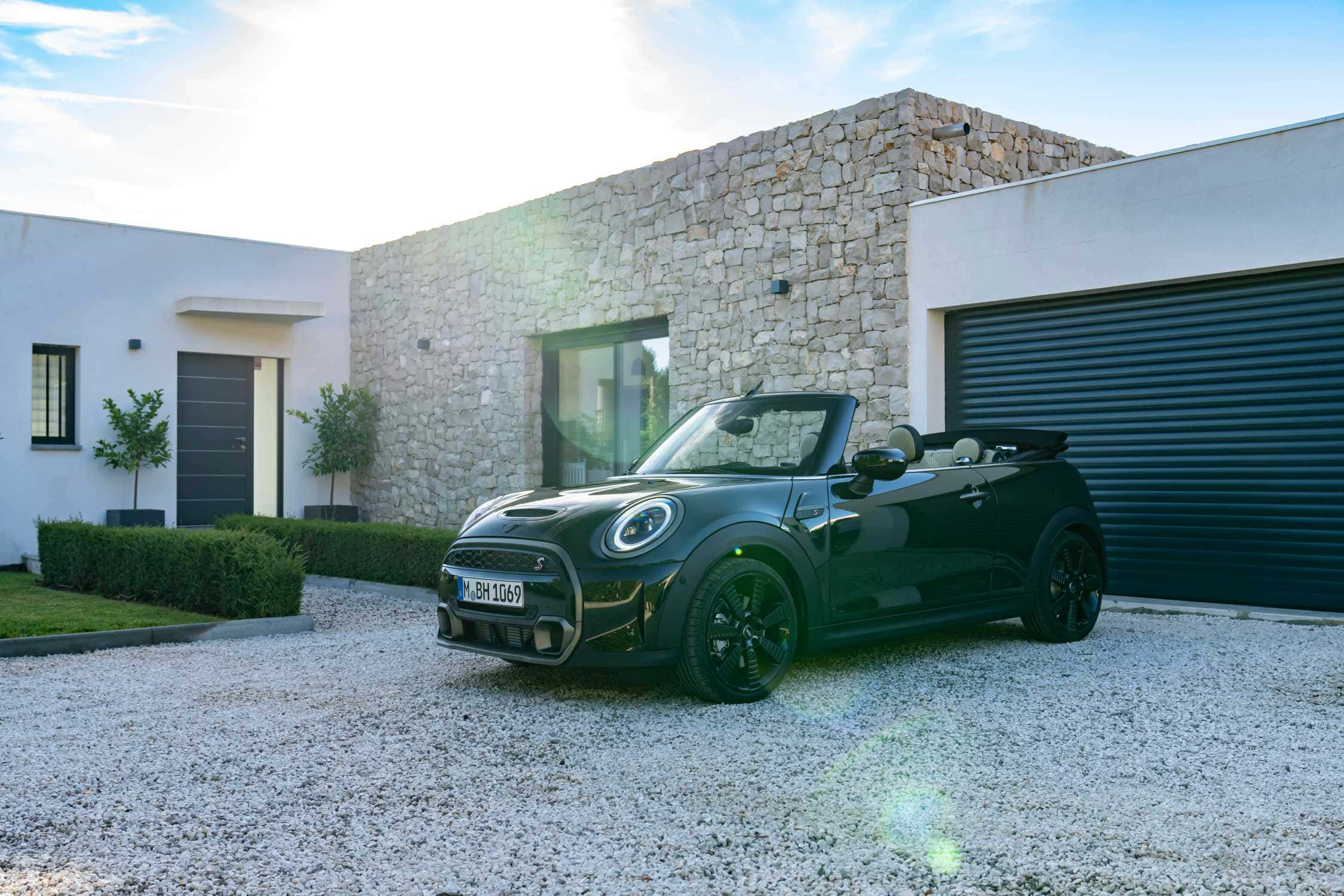 Mini Cooper S Convertible 桌布 壁紙 — 一架時尚的黑色MINI開篷車停在碎石車道上，前面是現代石頭和白色房屋，帶有太陽眩光。