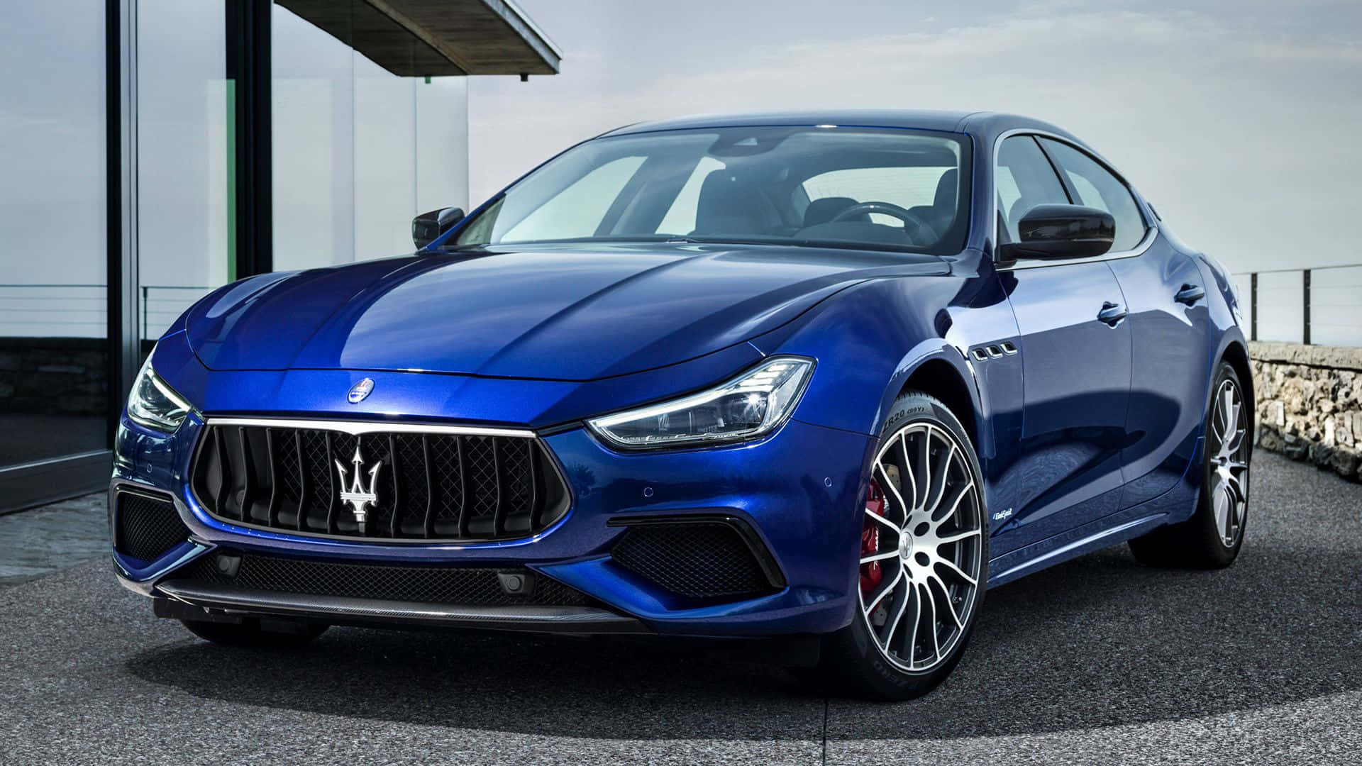 Maserati Ghibli 桌布 壁紙 — 一架時尚的藍色瑪莎拉蒂 Ghibli 跑車停泊在玻璃建築旁的現代石頭庭院上。