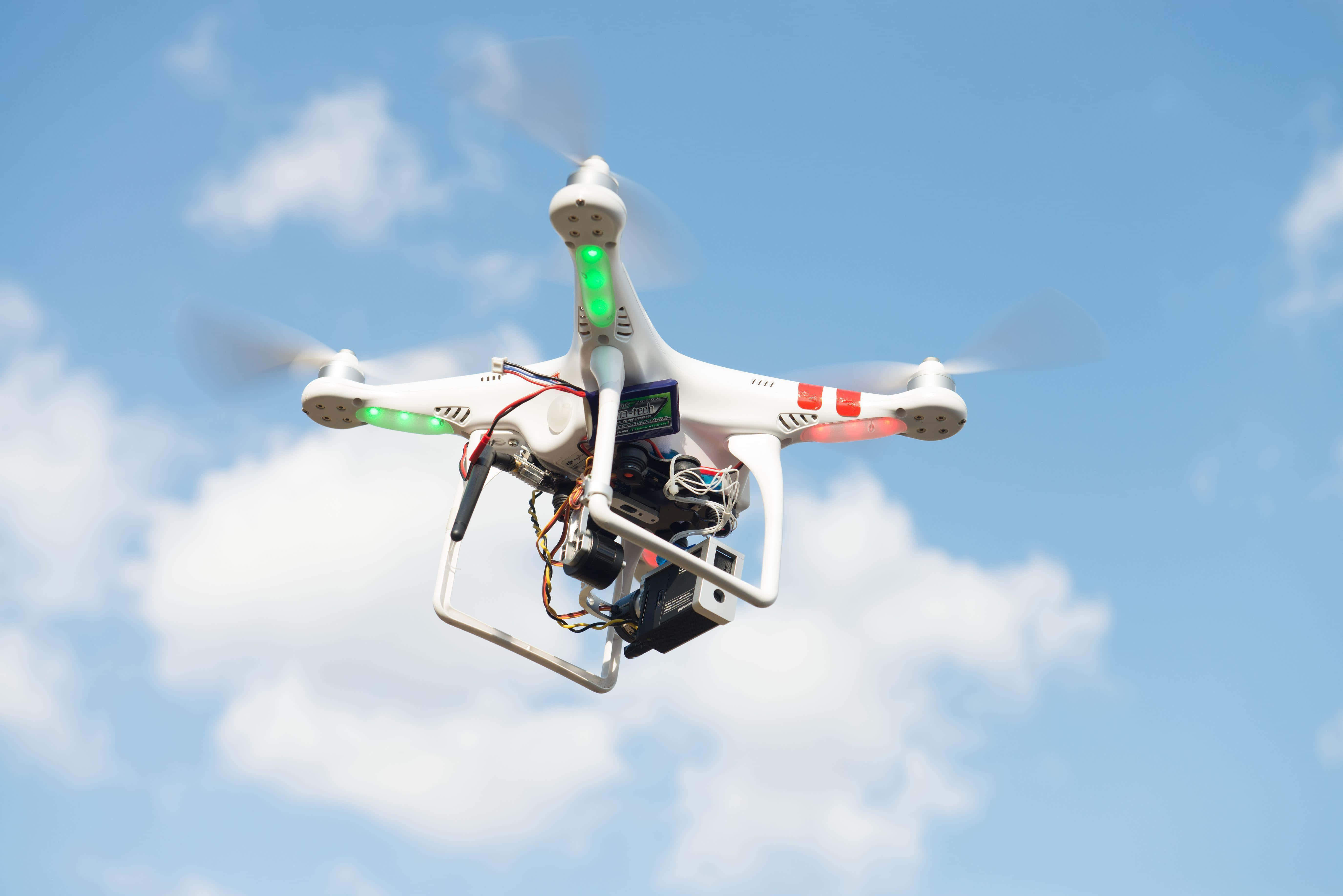Drone Camera 桌布 壁紙 — 一架白色四軸無人機在明亮的藍天和蓬鬆的白雲中盤旋，其螺旋槳快速旋轉。