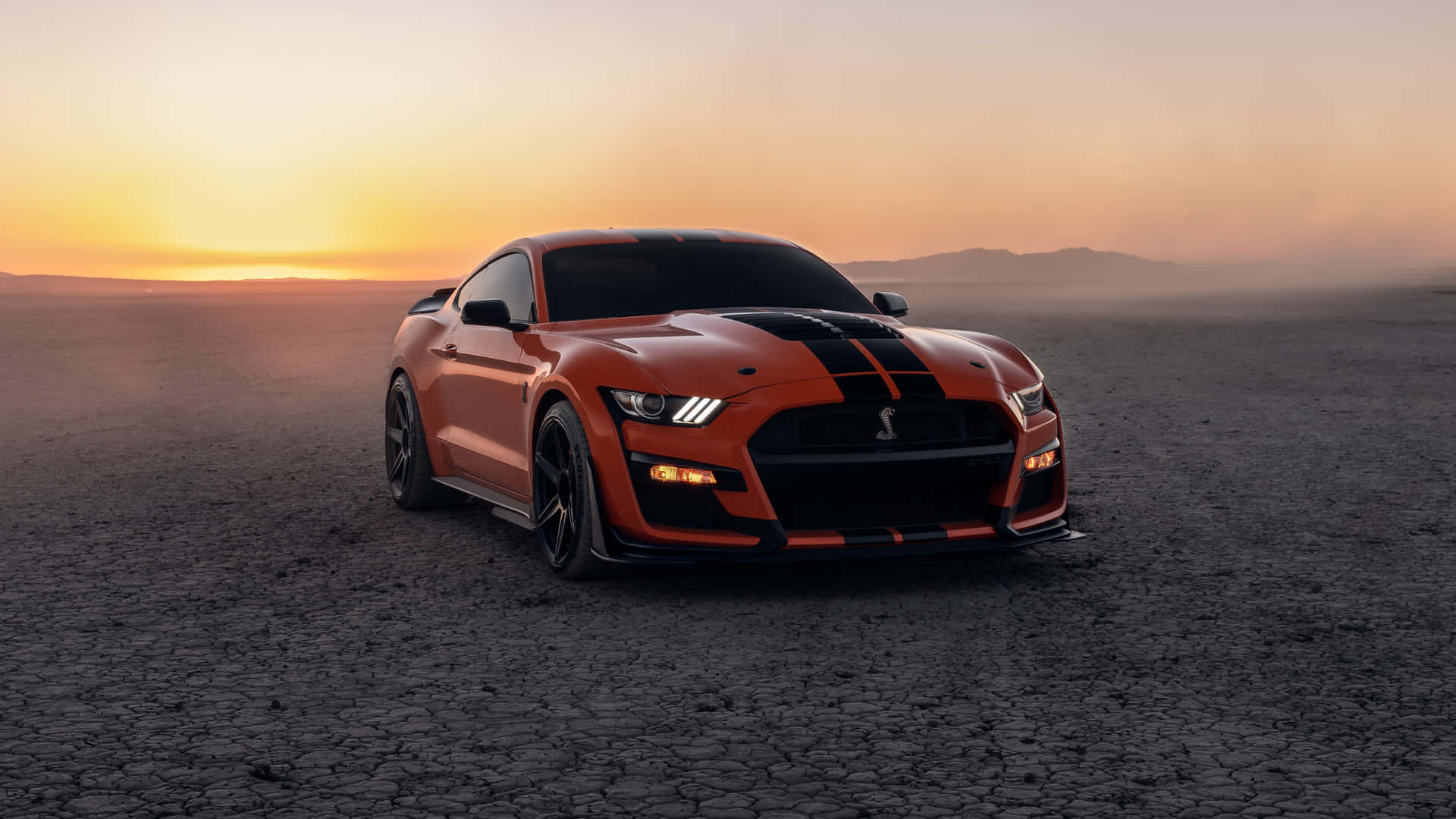  壁紙 — 一架帶有黑色賽車條紋的火橙色福特野馬Shelby GT500在日落時分立於龜裂的沙漠土地上。