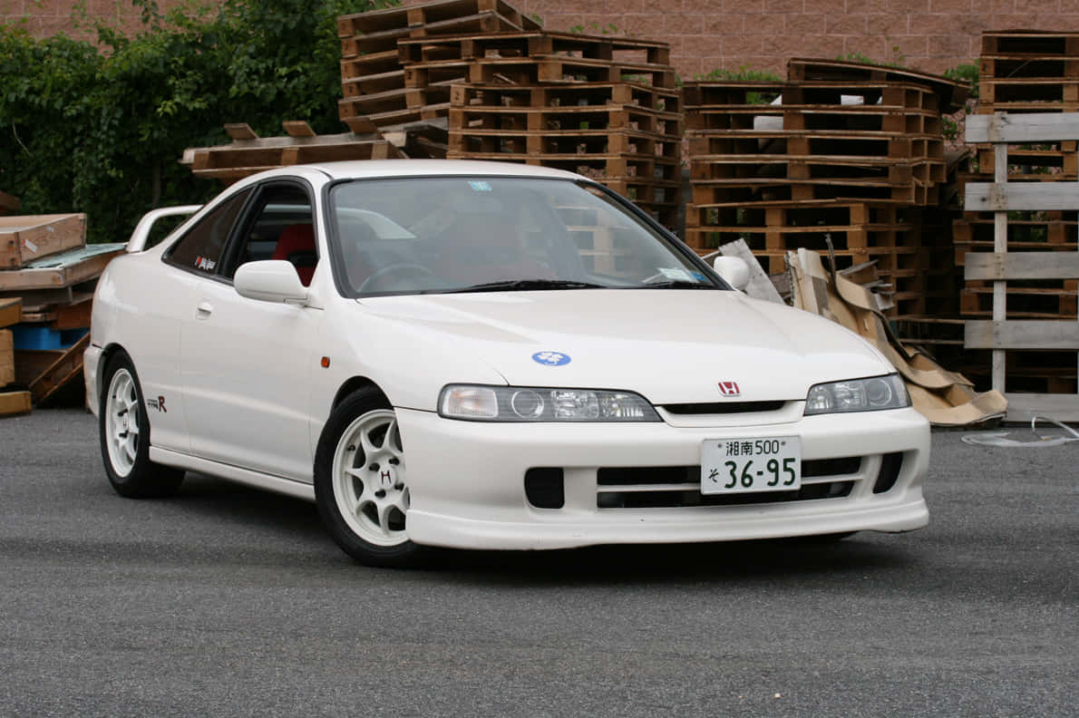 Honda Integra 桌布 壁紙 — 一架純白色本田Integra Type R雙門跑車停泊在瀝青路上，背景有堆疊的木托盤和綠植。