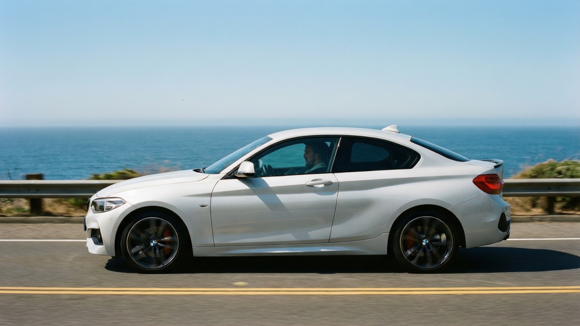 Bmw 2 Series 桌布 壁紙 — 一架時尚的白色BMW雙門轎跑車沿著海岸公路行駛，海洋在晴朗的藍天下延伸至地平線。