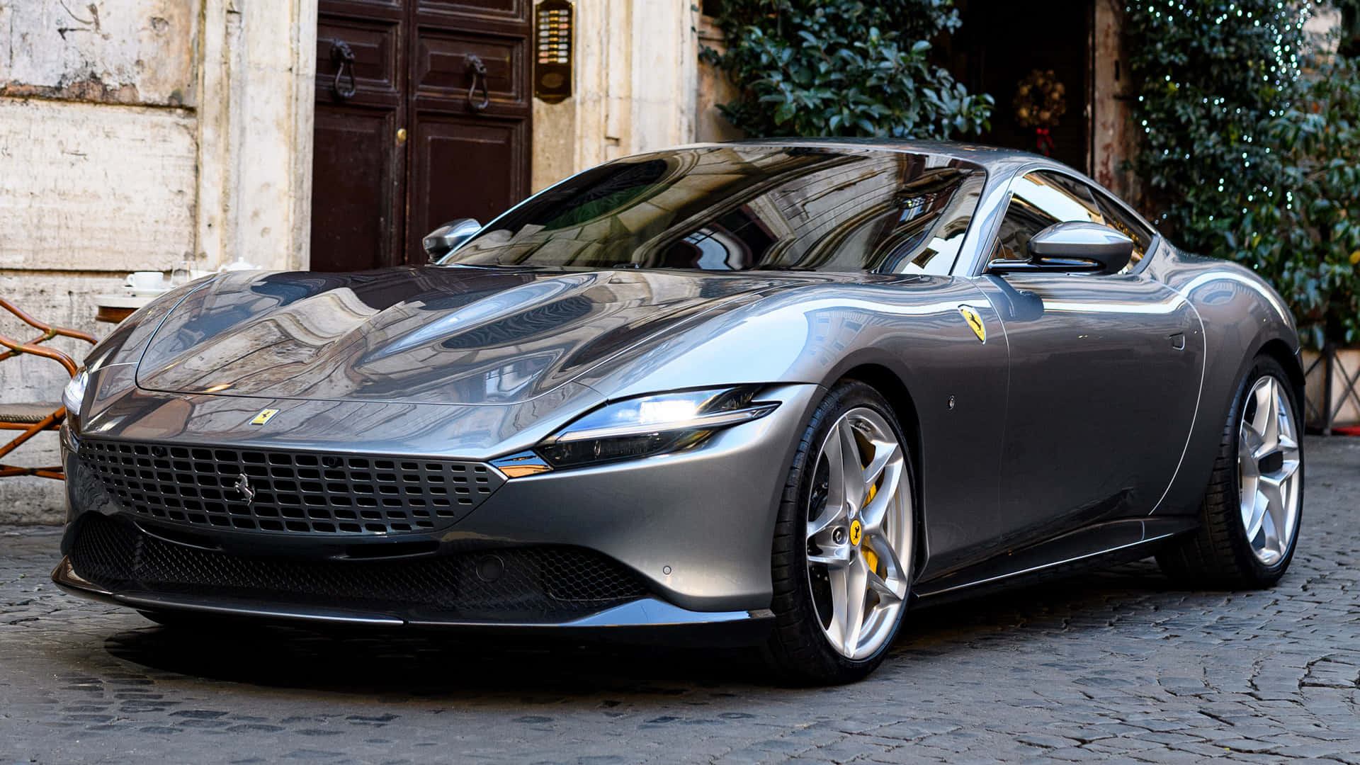 Ferrari Roma 桌布 壁紙 — 一架時尚的銀色法拉利停泊在鵝卵石街道上，旁邊是帶有木門和綠植的歷史建築。