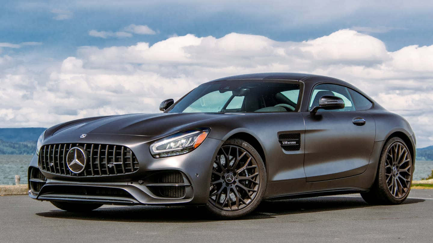 Mercedes Benz Amg Gt 桌布 壁紙 — 一架時尚的啞光灰色梅賽德斯-AMG GT跑車停泊在雲層密佈的天空下的沿海道路上。