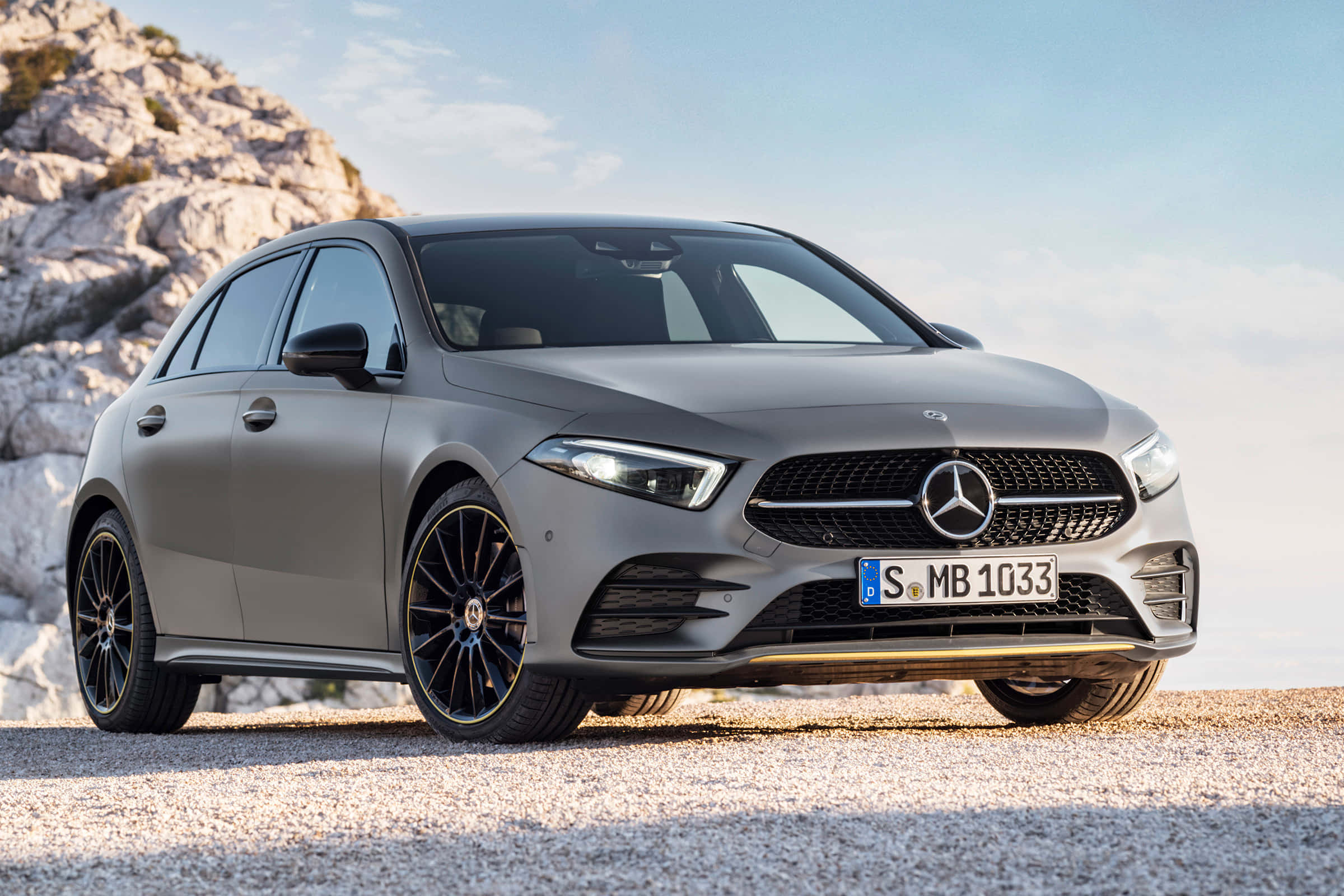 Mercedes Benz A-class 桌布 壁紙 — 一架時尚的啞光灰色平治A-Class停泊在礫石上，旁邊是岩石峭壁，上方是晴朗的藍天。