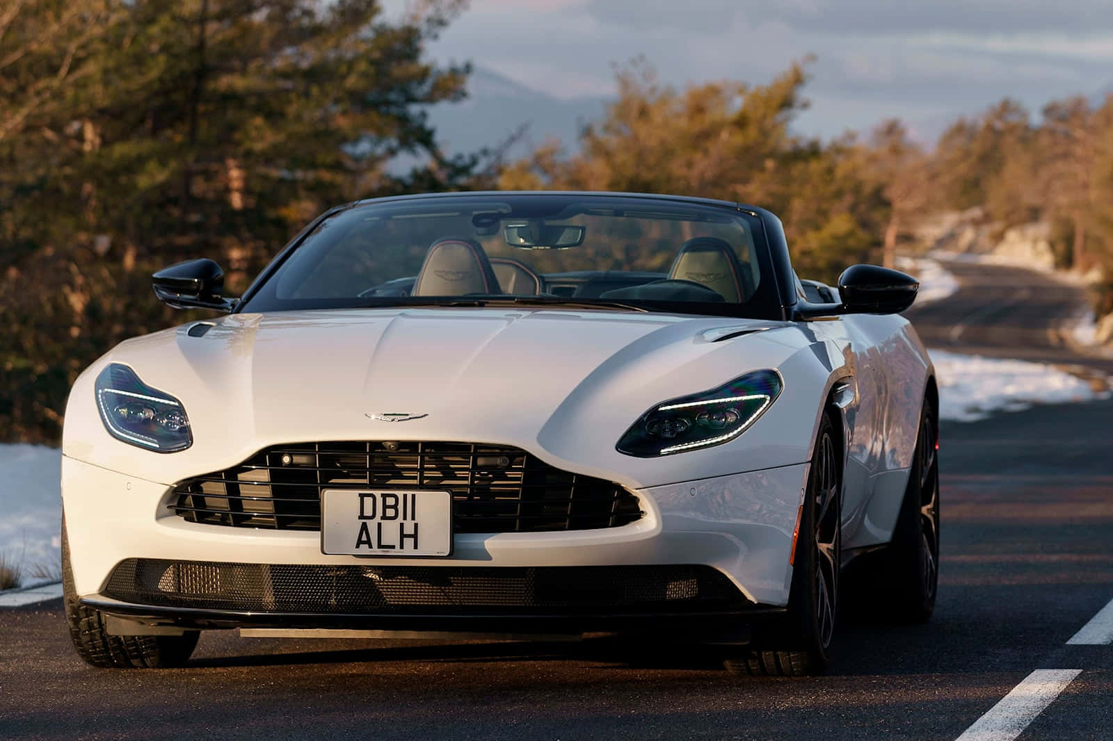 Aston Martin Db11 桌布 壁紙 — 一架時尚的白色Aston Martin DB11開篷跑車停泊在蜿蜒的山路上，周圍有被雪覆蓋的樹木和遠峰，背景是柔和的金色日落。