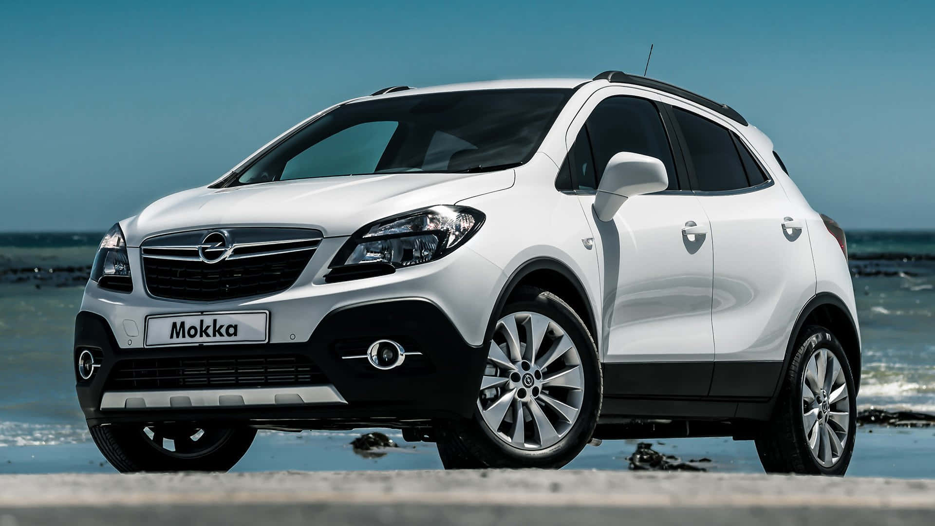 Opel Mokka 桌布 壁紙 — 一架時尚的白色歐寶 Mokka SUV 停泊在海岸混凝土板上，背後有海浪拍打，天空湛藍清澈。