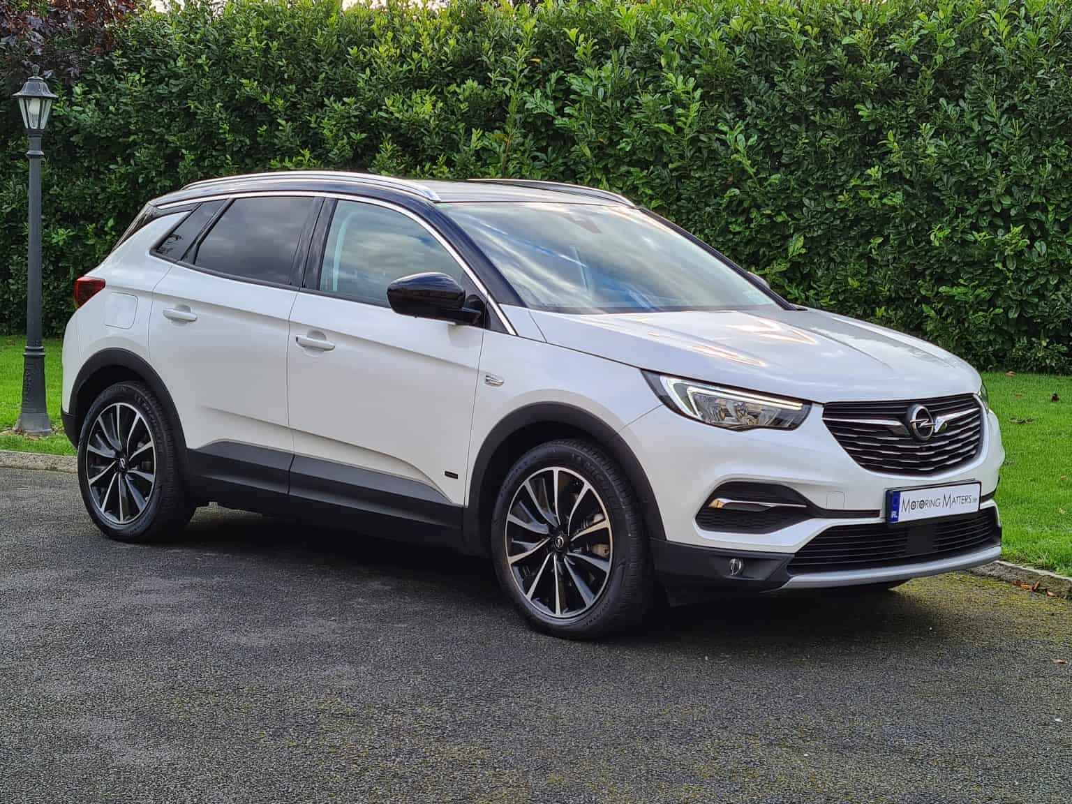 Opel Grandland X 桌布 壁紙 — 一架時尚的白色Opel SUV停泊在鋪砌的車道上，旁邊是茂密的綠色籬笆和街燈柱。
