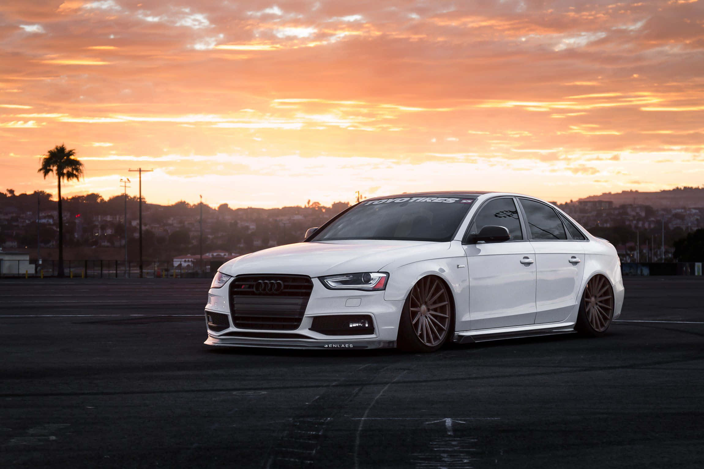 Audi S4 桌布 壁紙 — 一架時尚的白色奧迪轎車，配備定制青銅輪圈，停在瀝青路面上，背景是火紅的橙色日落天空。