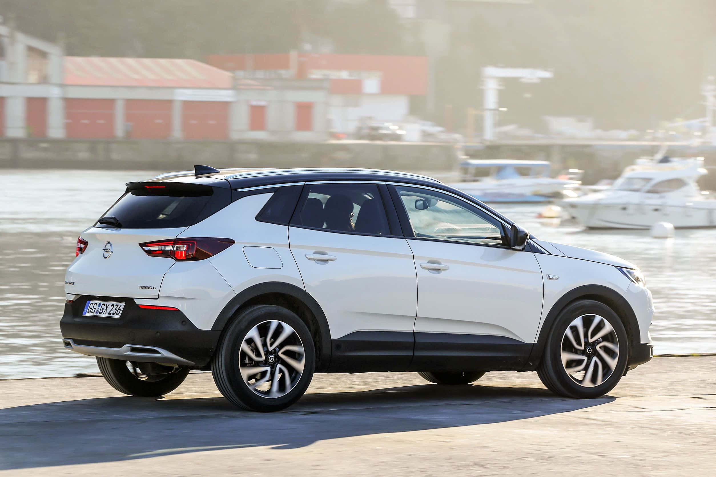Opel Grandland X 桌布 壁紙 — 一架時尚的白色SUV停泊在港口，背景有船隻和紅色建築物，略顯朦朧。