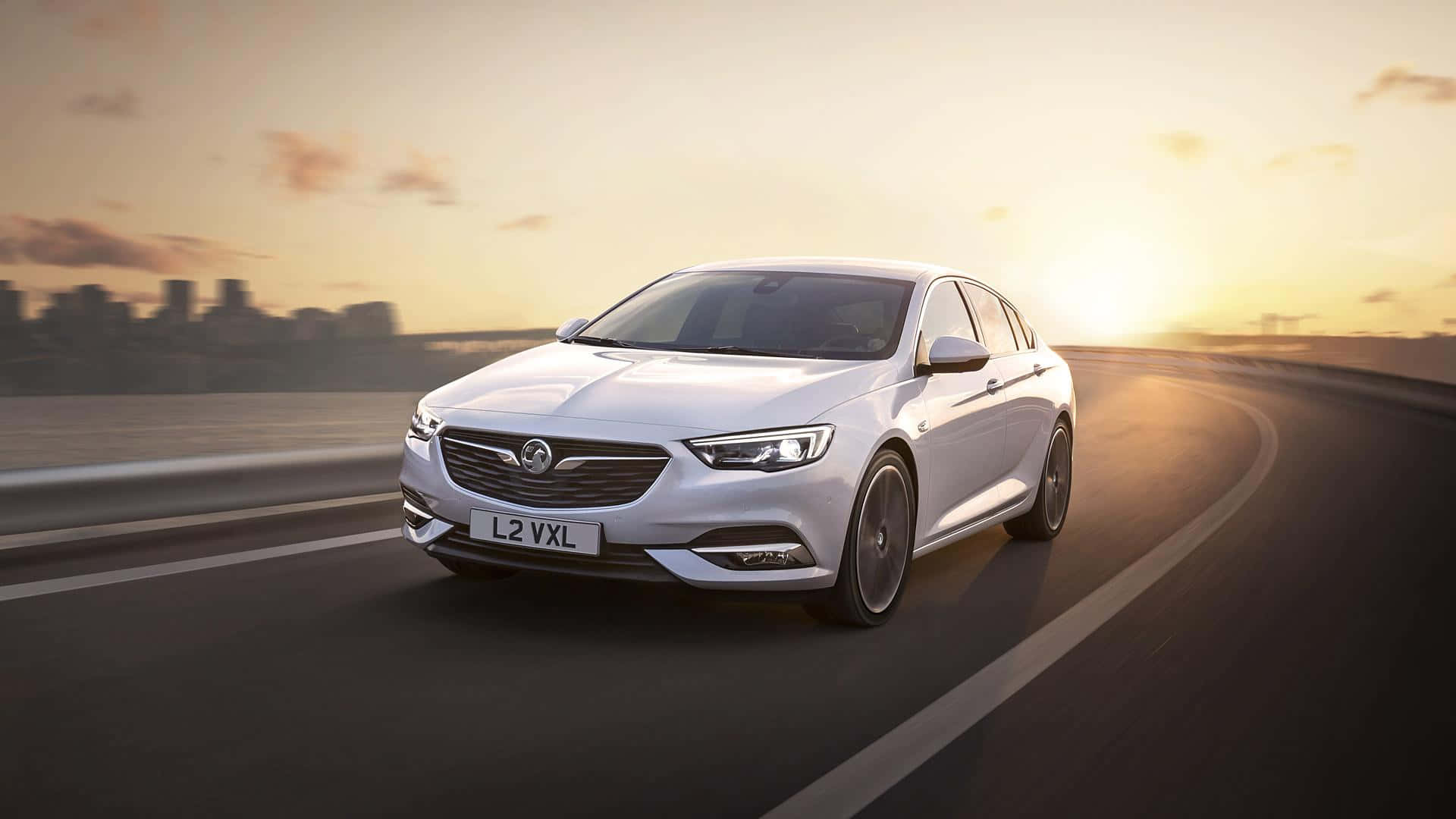 Opel Insignia 桌布 壁紙 — 一架時尚的白色Vauxhall房車在日落時分沿著彎曲的高速公路飛馳，遠處是模糊的城市天際線。