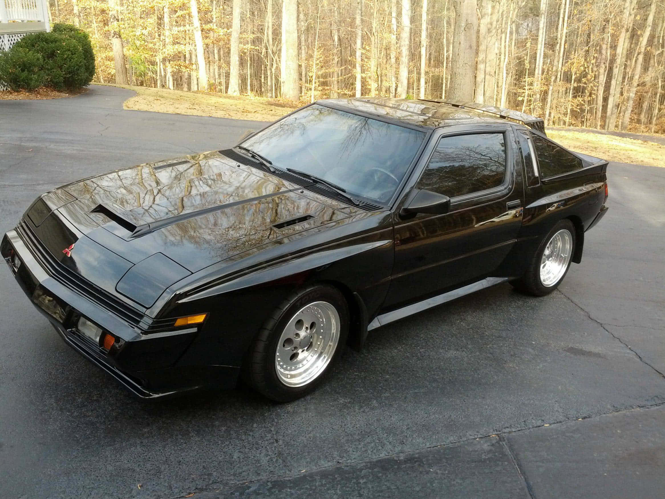 Mitsubishi Starion 桌布 壁紙 — 一架時尚的黑色三菱Starion跑車停泊在車道上，光亮的引擎蓋反射出光禿禿的樹木。