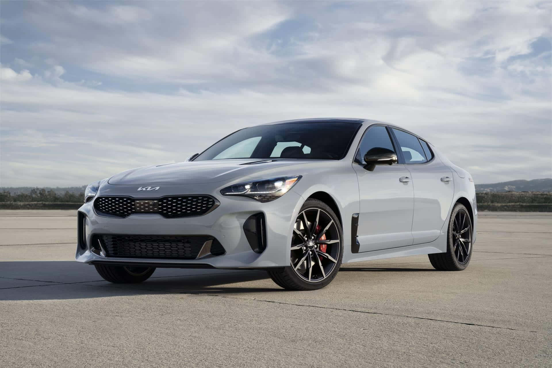 Kia Stinger 桌布 壁紙 — 一架時尚的銀色Kia Stinger運動轎車，帶有黑色飾件和紅色制動卡鉗，停放在混凝土停車場上，天空多雲。
