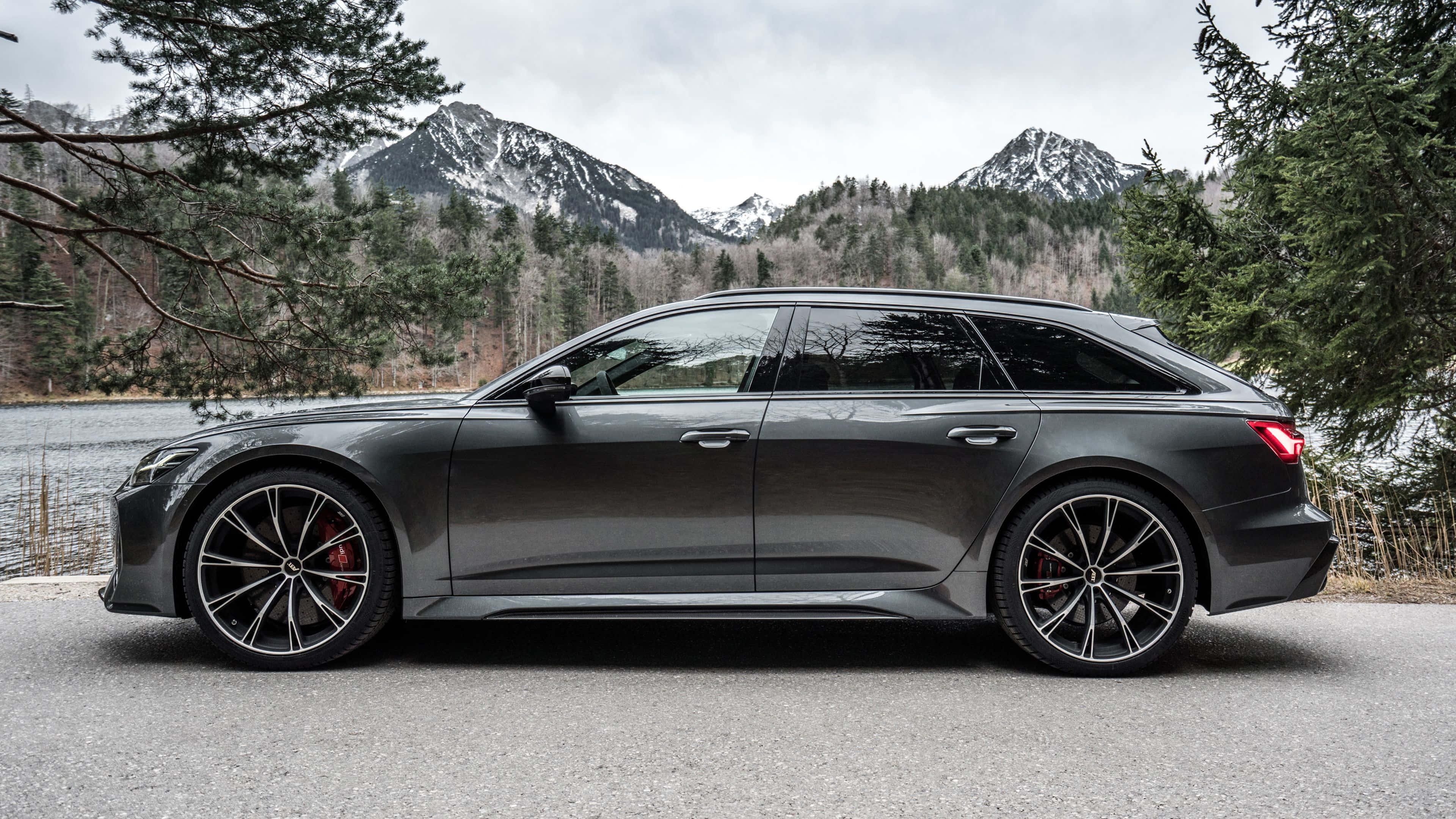 Audi Rs6 桌布 壁紙 — 一架時尚的深灰色奧迪旅行車停泊在湖邊，背景是白雪覆蓋的山脈和常綠樹木。