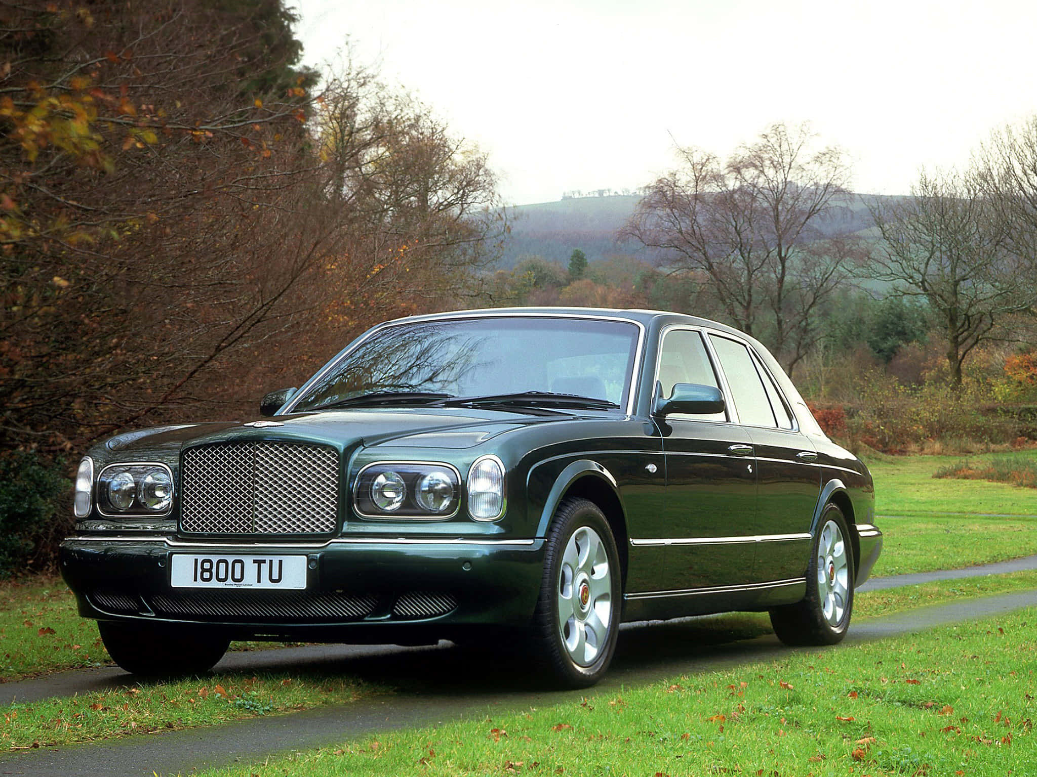 Bentley Arnage 桌布 壁紙 — 一架時尚的深綠色賓利 Arnage 停泊在蜿蜒的道路上，周圍環繞著秋天的樹木和起伏的山丘。