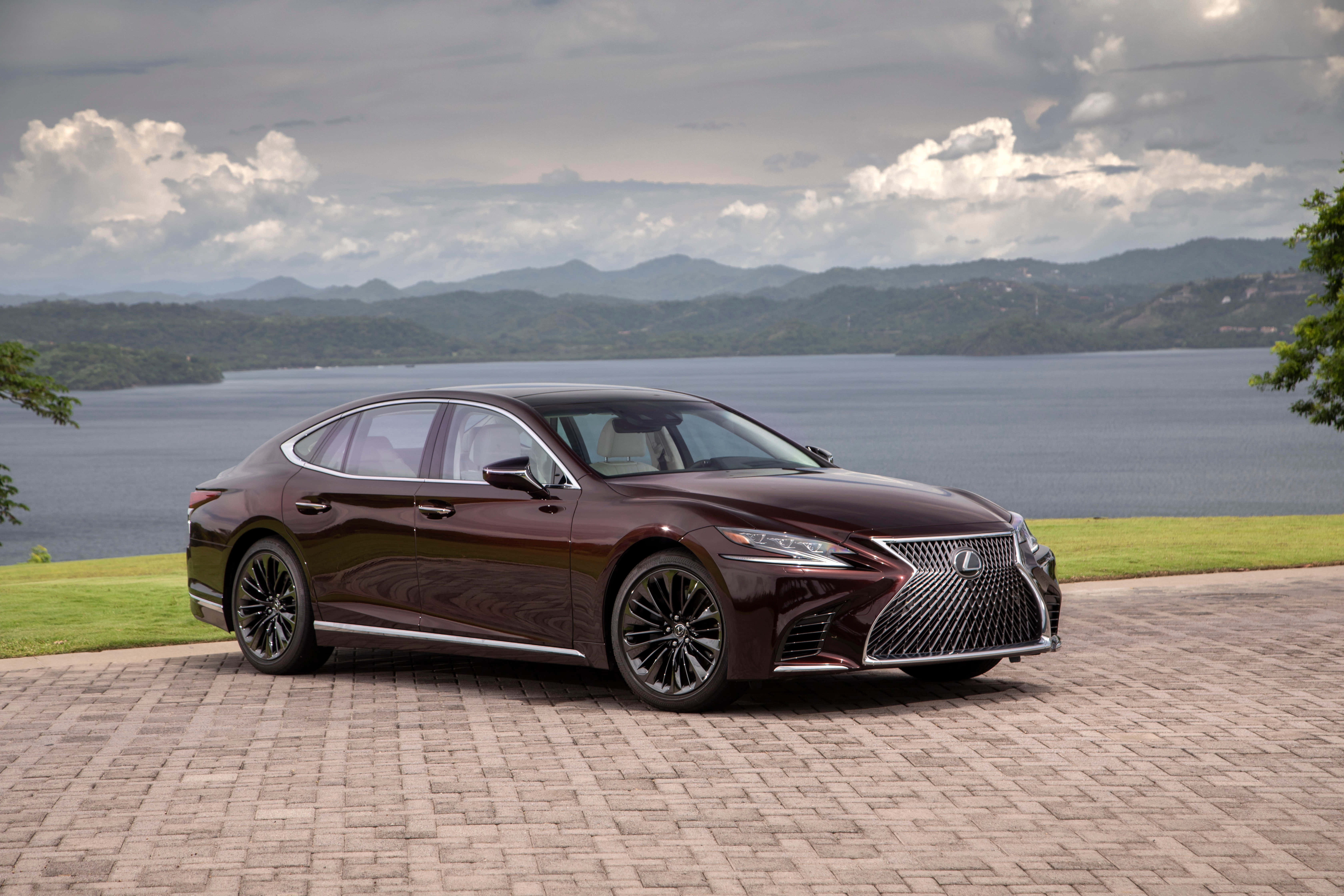 Lexus Ls 500 桌布 壁紙 — 一架時尚的酒紅色凌志房車停泊在石頭車道上，俯瞰廣闊的湖泊，遠處有起伏的山丘和陰天。
