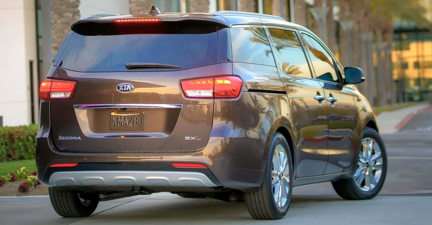Kia Sedona 桌布 壁紙 — 一架時尚的棕色Kia Sedona小型貨車停泊在陽光明媚的街道上，背景有棕櫚樹和建築物。