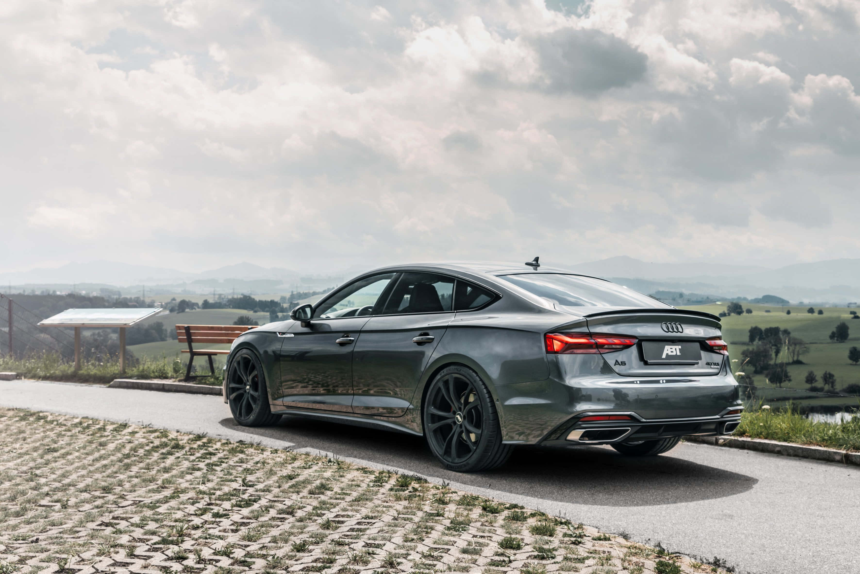 Audi A5 桌布 壁紙 — 一架時尚灰色ABT Audi A5 Sportback停泊在鵝卵石路上，俯瞰著多雲天空下連綿起伏的綠色山丘。