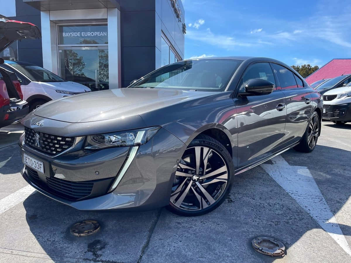 Peugeot 508 桌布 壁紙 — 一架時尚金屬灰色標緻508房車停泊在陽光普照的經銷商場地，背景有其他汽車和現代建築。