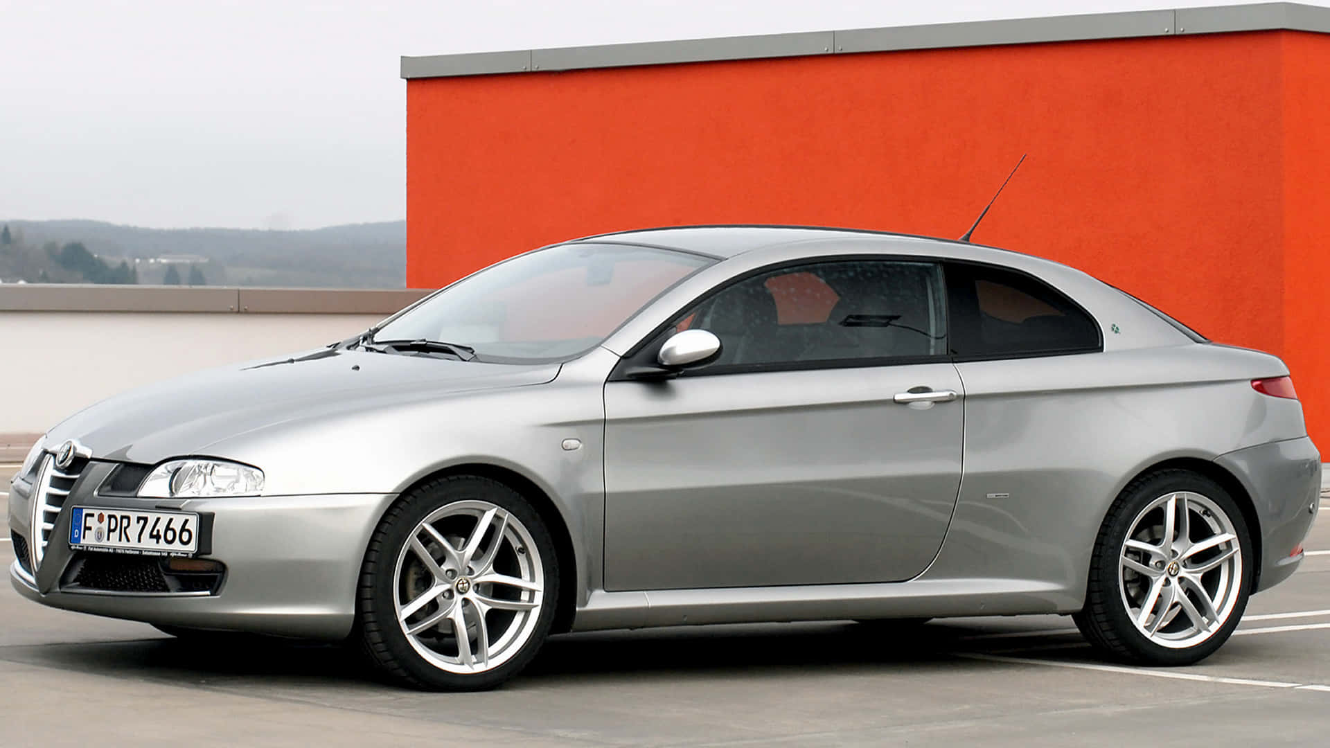 Alfa Romeo Gt 桌布 壁紙 — 一架時尚的銀色愛快羅密歐雙門跑車停泊在陰天下一道大膽的橙色牆旁邊。