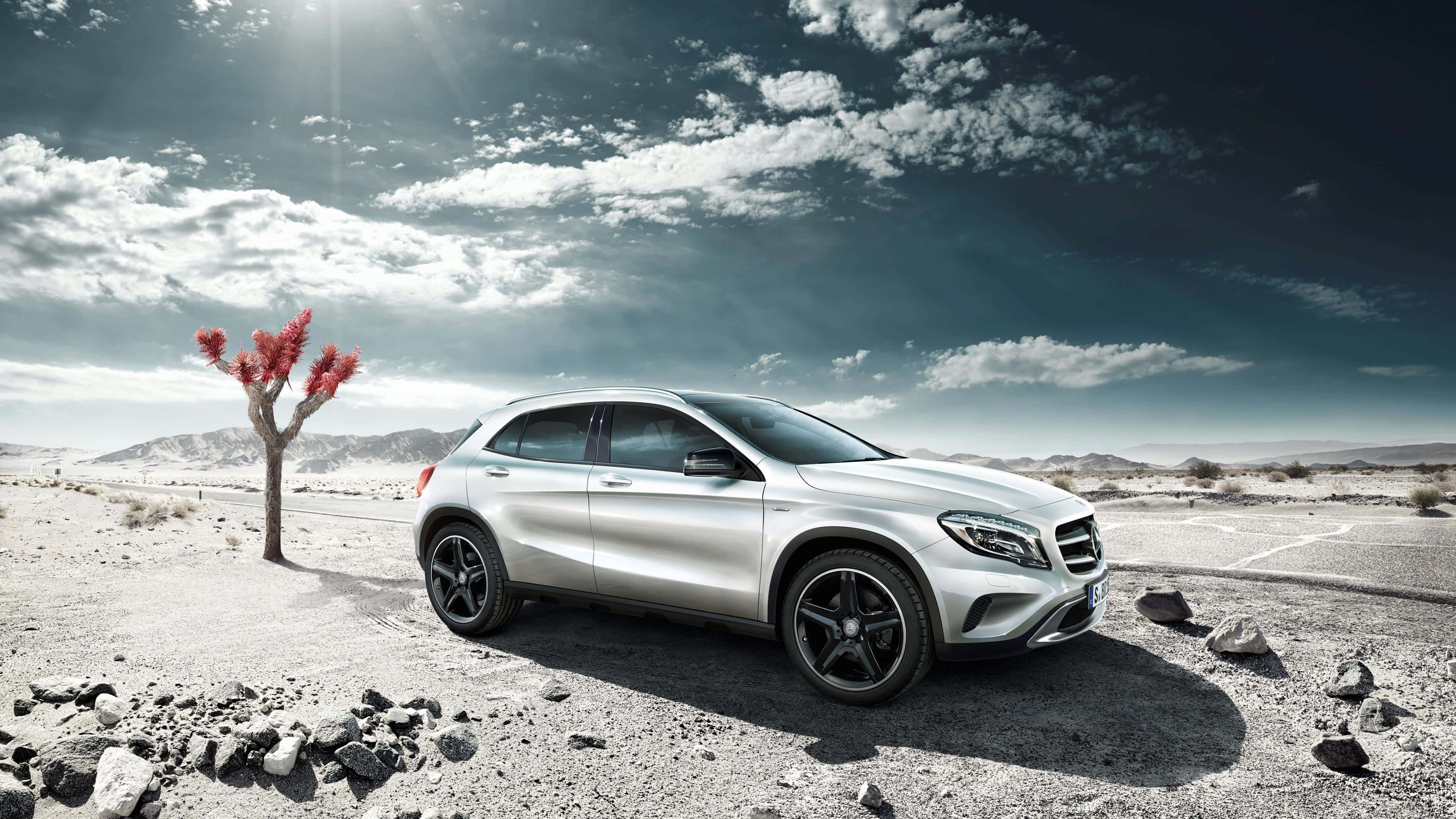 Mercedes Benz Gla-class 桌布 壁紙 — 一架時尚的銀色SUV停泊在沙漠公路上，旁邊有一棵孤獨的紅葉樹，天空佈滿戲劇性的雲層。