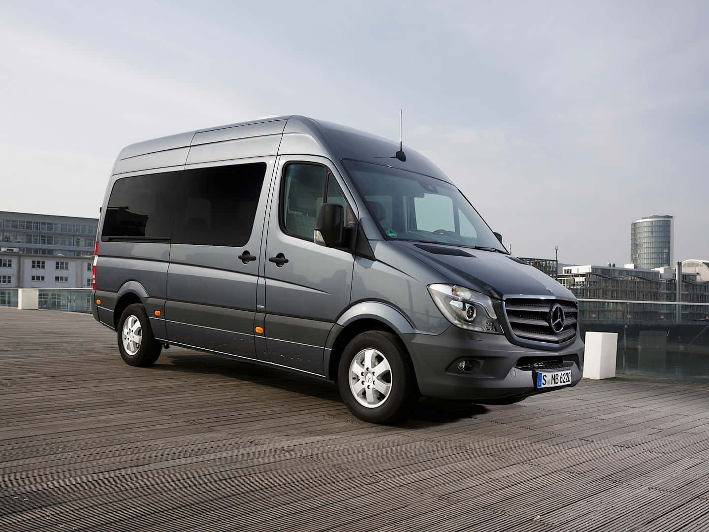 Mercedes Benz Sprinter 桌布 壁紙 — 一架時尚的灰色 Mercedes-Benz Sprinter 客貨車停泊在木製屋頂上，背景是現代建築物，天空多雲。