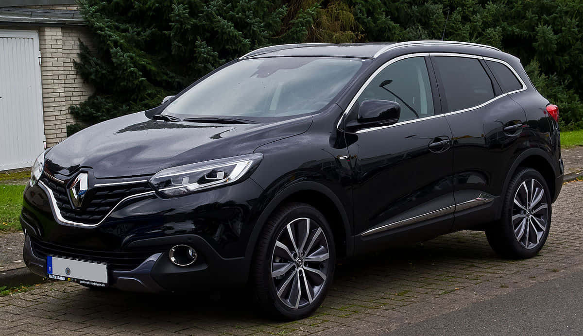  壁紙 — 一架時尚的黑色雷諾 Koleos SUV 停泊在磚屋旁邊的鵝卵石車道上，背景有綠化植物。
