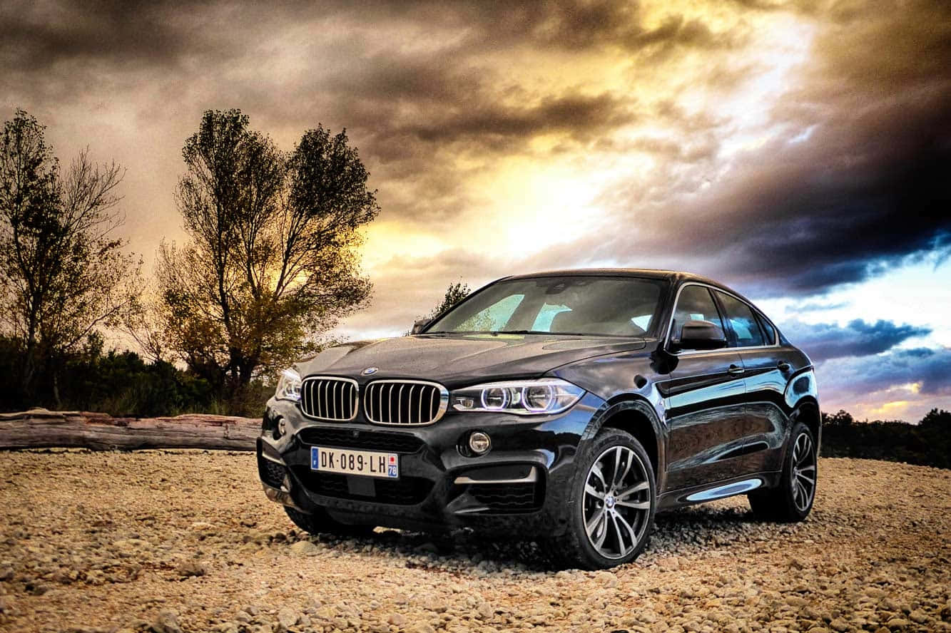 Bmw X6 桌布 壁紙 — 一架時尚黑色BMW X6 SUV停泊在碎石場上，背景是戲劇性的日落天空，金色雲朵和樹木剪影。