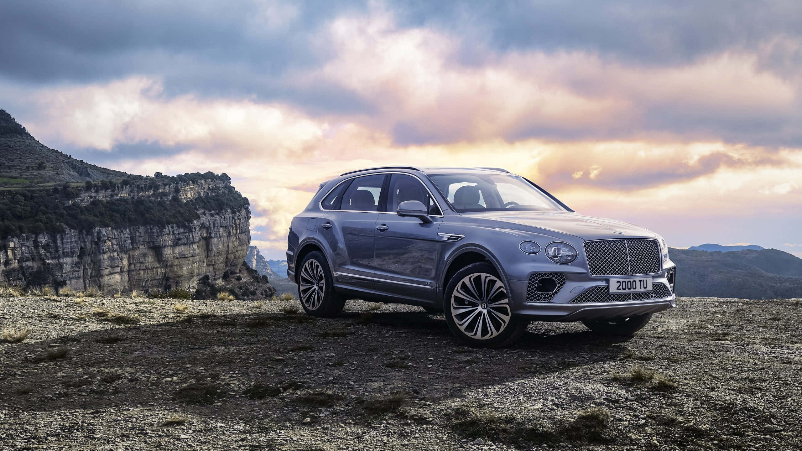 Bentley Bentayga 桌布 壁紙 — 一架時尚的銀色賓利Bentayga SUV在日落時分矗立在崎嶇的懸崖上，背後是戲劇性的雲層和岩石地形。