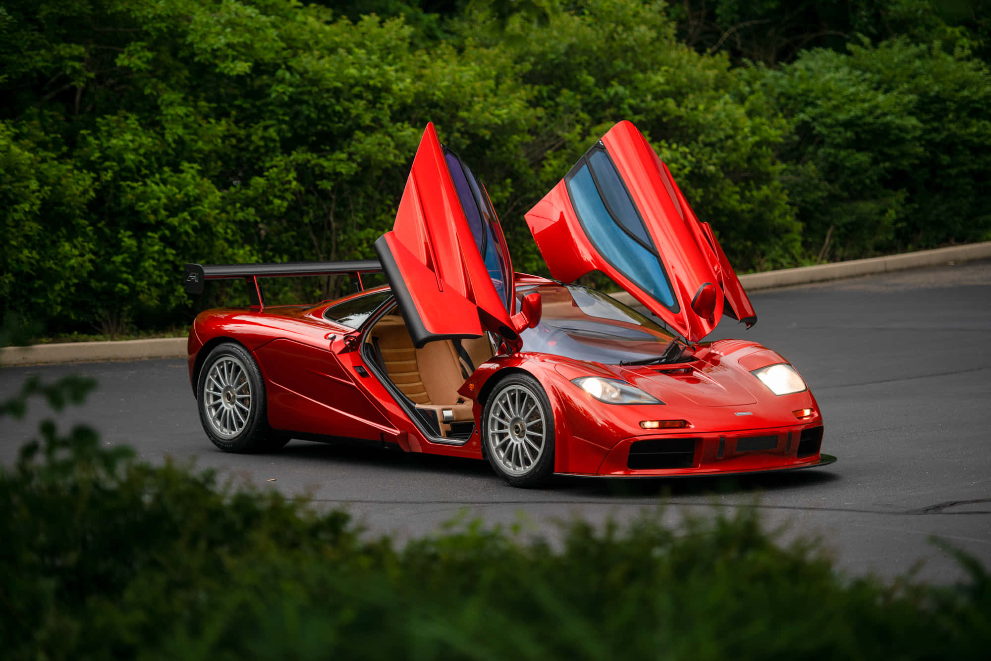 Mclaren F1 桌布 壁紙 — 一架引人注目的紅色McLaren F1超級跑車，鷗翼車門打開，停泊在綠樹成蔭的道路上。