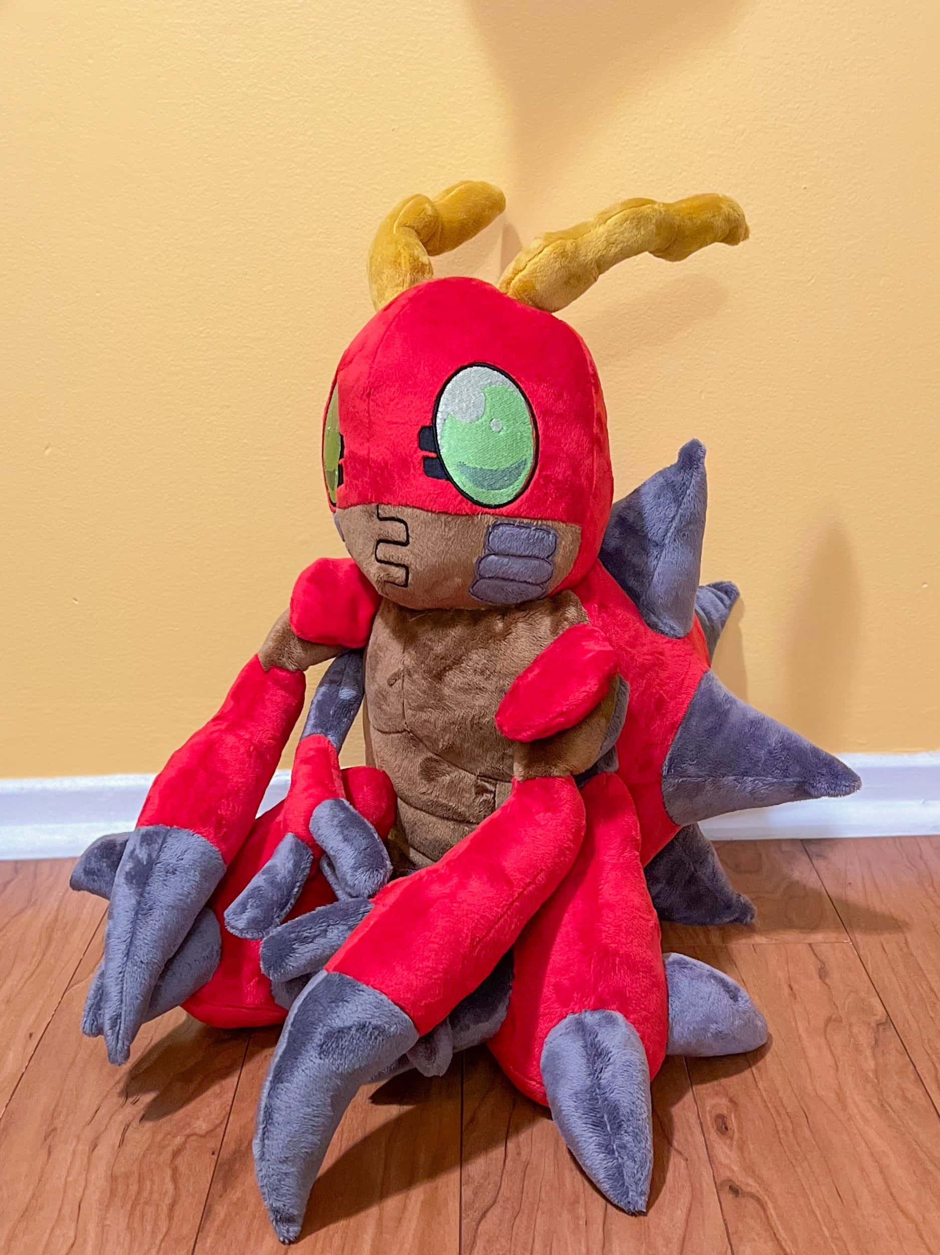 Digimon Tentomon 桌布 壁紙 — 一個大型紅色和灰色毛絨玩具，有綠色眼睛和黃色角，坐在木地板上，背靠黃色牆壁。