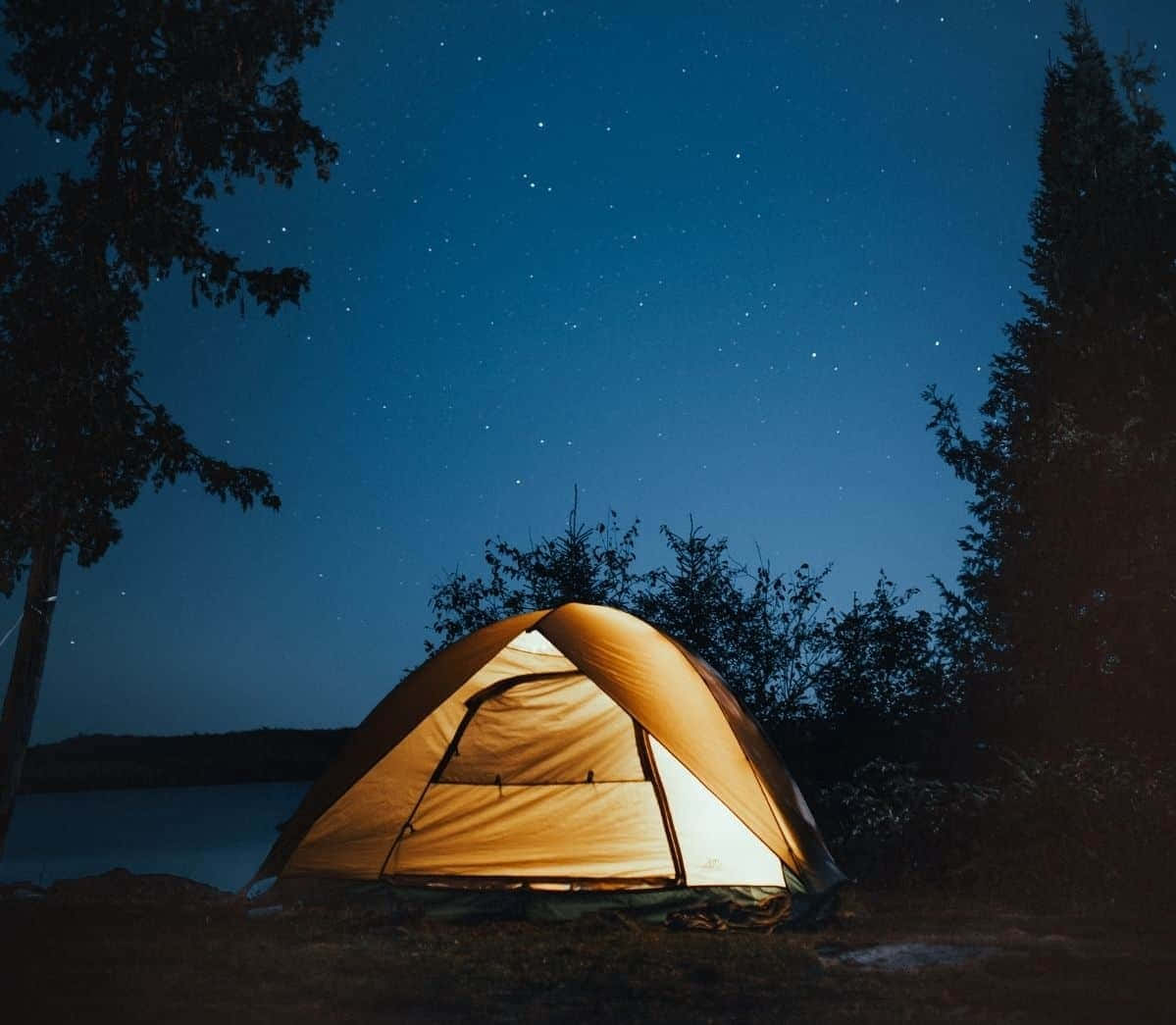 在繁星點點的夜空下，湖邊一頂溫暖照亮的帳篷，周圍樹木環繞。