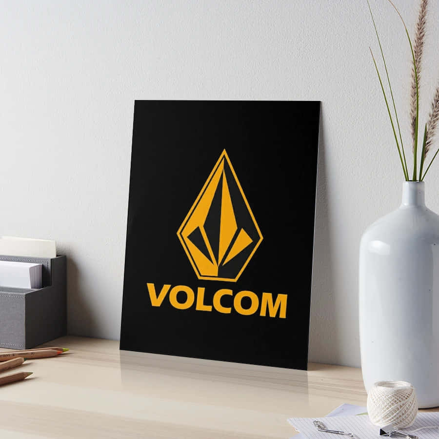  壁紙 — 一個黑色Volcom標誌藝術印刷品，帶有鮮明的黃色品牌標識，靠在一張木桌旁的白牆上，旁邊有一個花瓶和文具。