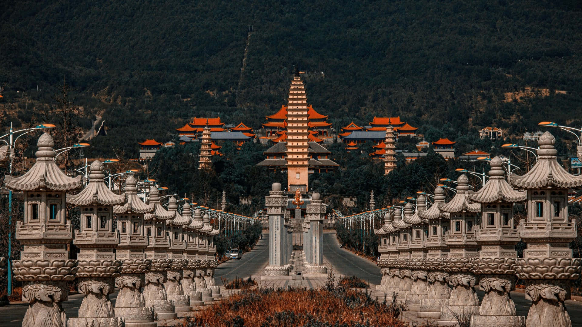  壁紙 — 一個宏偉的寺廟建築群，橙色屋頂和石燈籠排列在一條通往森林山丘中高聳寶塔的道路兩旁。
