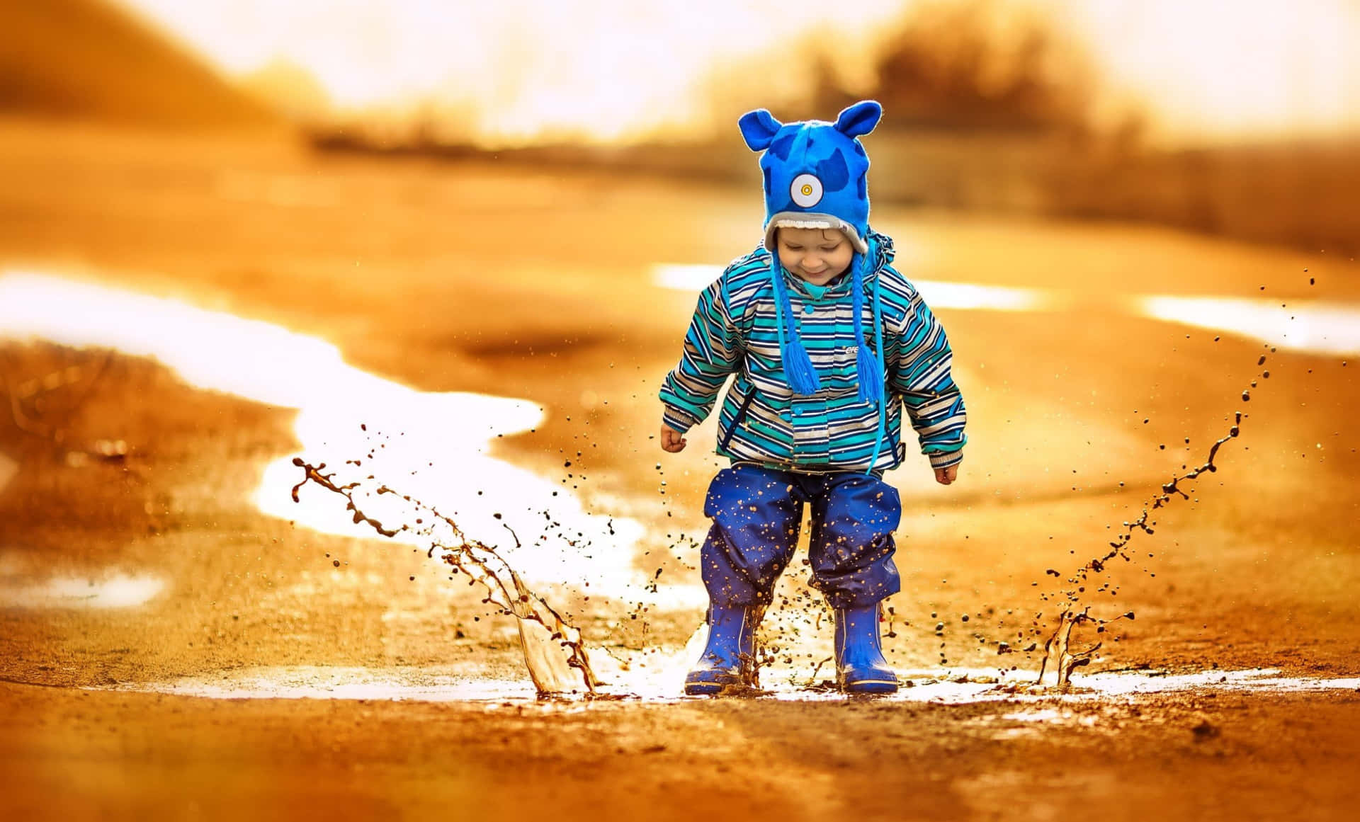  壁紙 — 一個快樂的幼兒戴著藍色動物帽和靴子在溫暖的金色陽光下踩過泥濘的水坑。