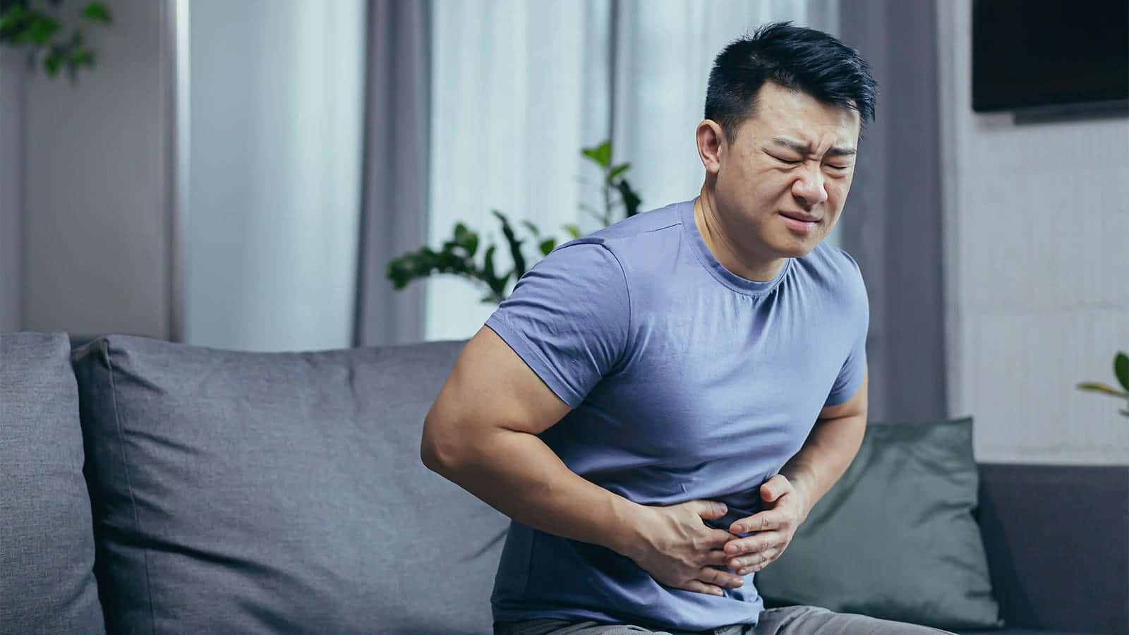  壁紙 — 一個男人坐在灰色梳化上，明顯痛苦地抓著肚子，臉上露出痛苦的表情。