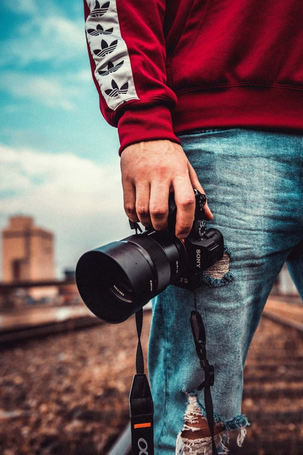 Cameraman 桌布 壁紙 — 一個人穿著紅色運動衫和破洞牛仔褲，在屋頂上拿著一部黑色Sony相機，背景是模糊的城市天際線。