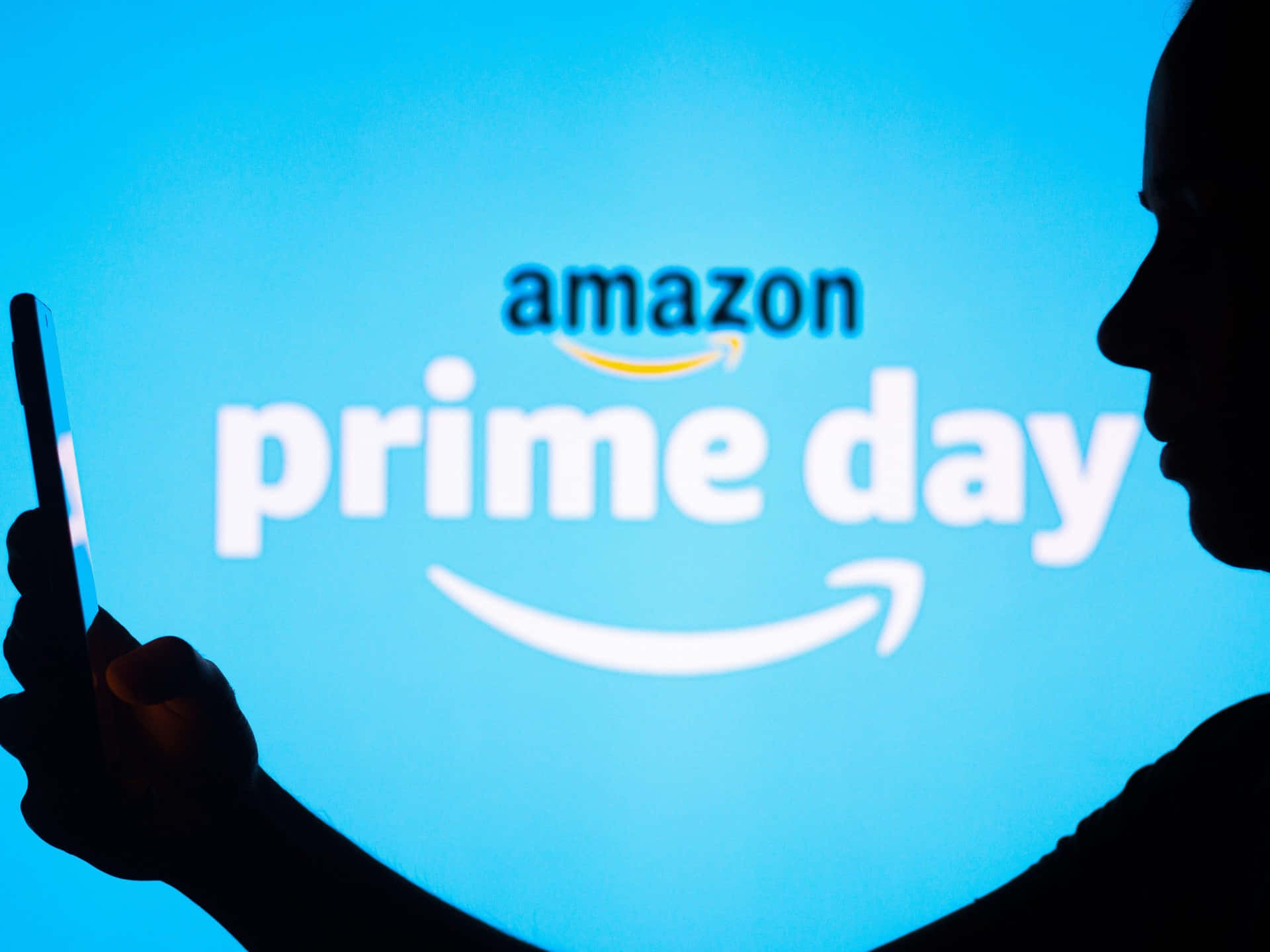  壁紙 — 一個人在發光的藍色Amazon Prime Day標誌前拿著手機的剪影。