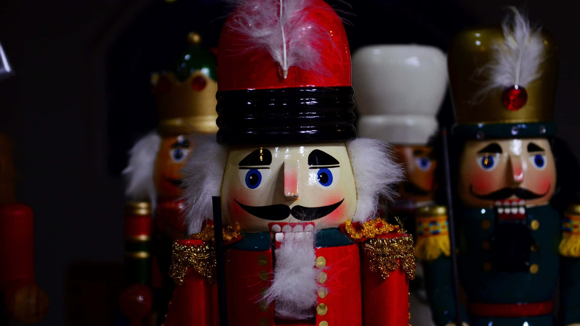  壁紙 — 一個閃亮紅色胡桃夾子，藍眼睛白鬍鬚，在昏暗燈光下與其他節日胡桃夾子站在一起的近距離特寫。