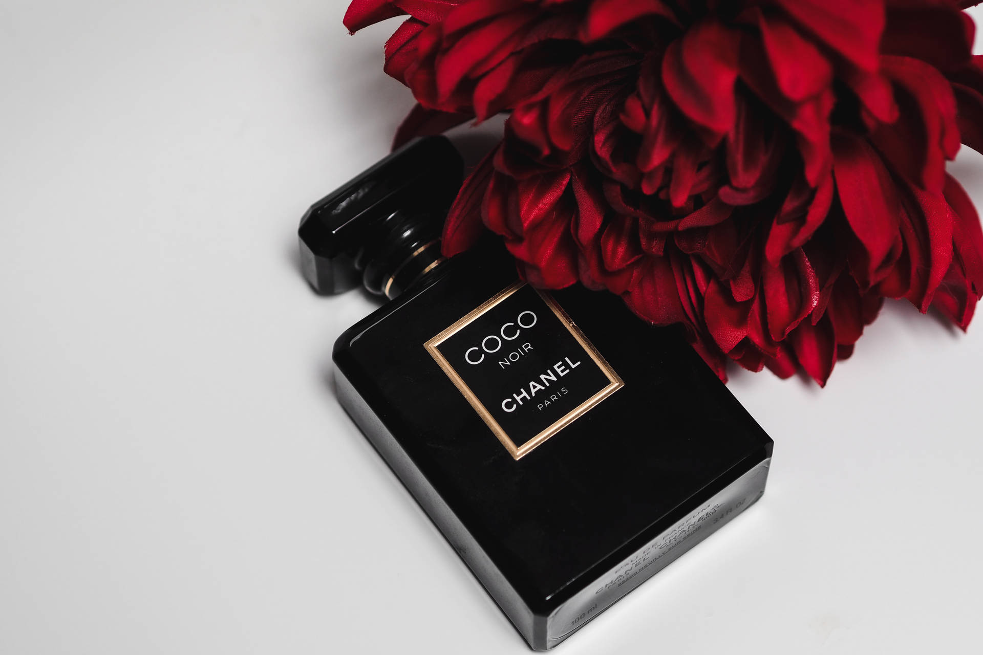 香奈兒Coco Noir紅玫瑰 壁紙 — 一個時尚的黑色 Chanel Coco Noir 香水瓶放在純白表面上，旁邊是一朵茂盛的紅花。