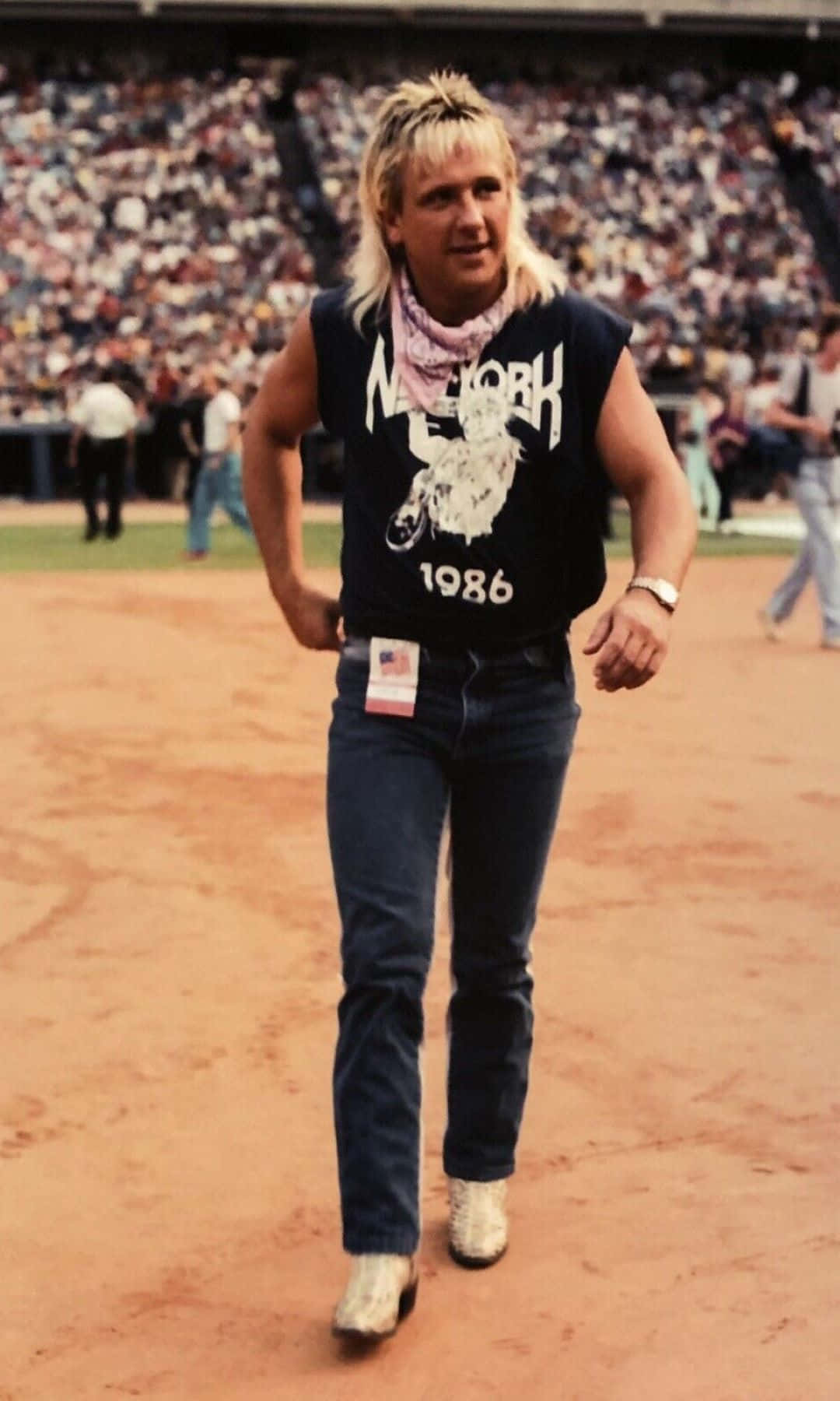  壁紙 — 一個頭髮尖尖的金髮男子自信地走在棒球場上，身穿1986年紐約圖案背心和牛仔褲。